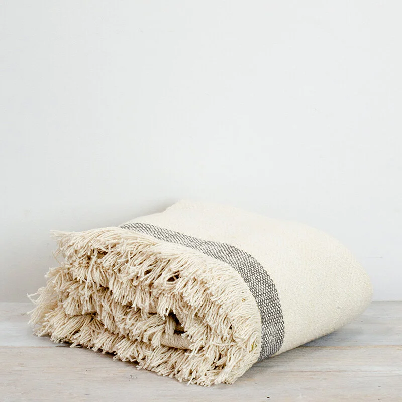 Folded beige throw blanket with fringe on a light wood surface.