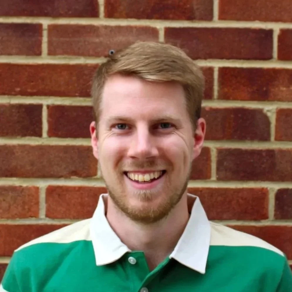 A white man with blonde hair and beard stands smiling in front of a brick wall. 