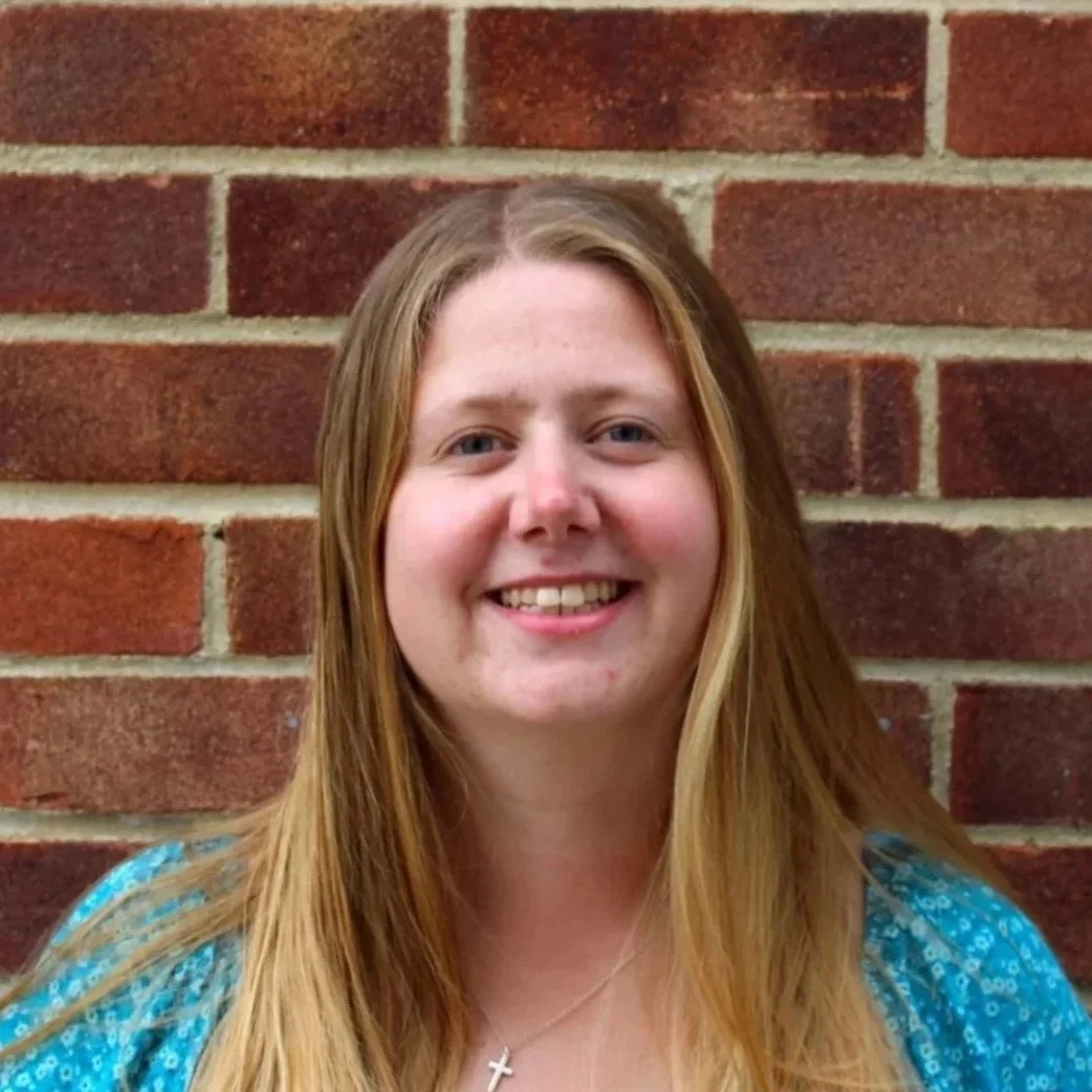 A white woman with blonde hair stands smiling in front of a brick wall. 