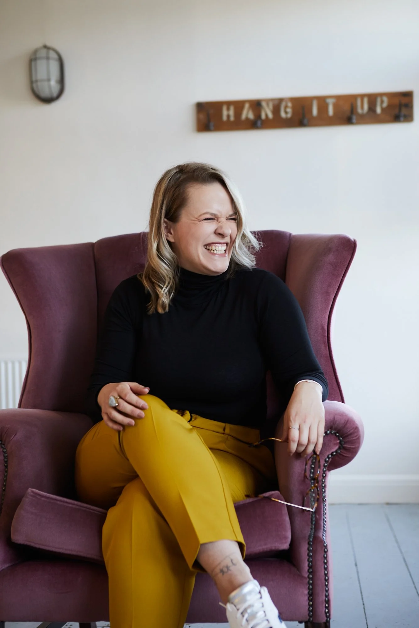 Cherish Amber sex and relationship coach sitting in pink velvet wingback chair laughing to the left.