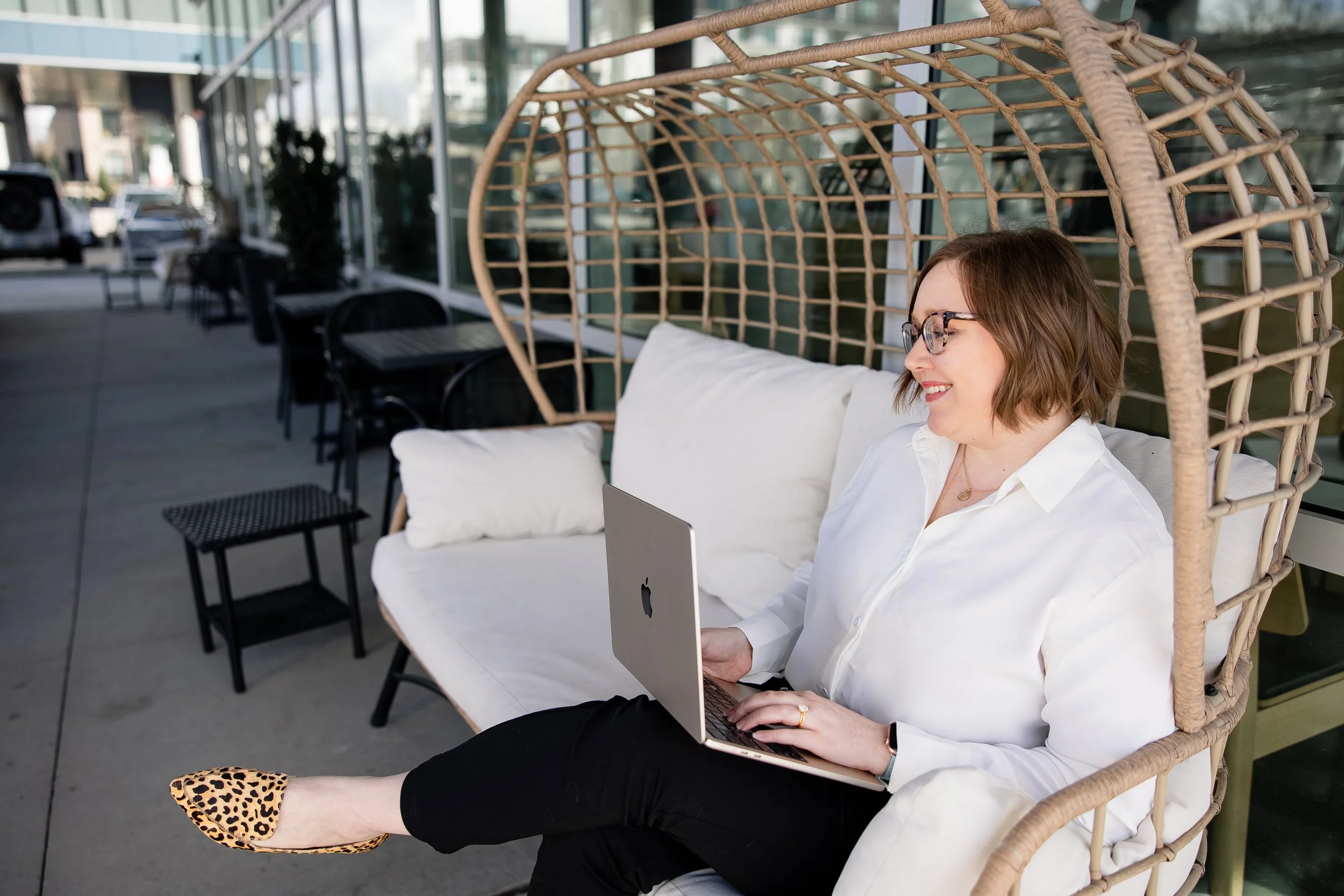 A woman sitting in a wicker chair outdoors, smiling, working on a laptop, wearing glasses, a white shirt, and leopard print shoes, next to a building with large glass windows.