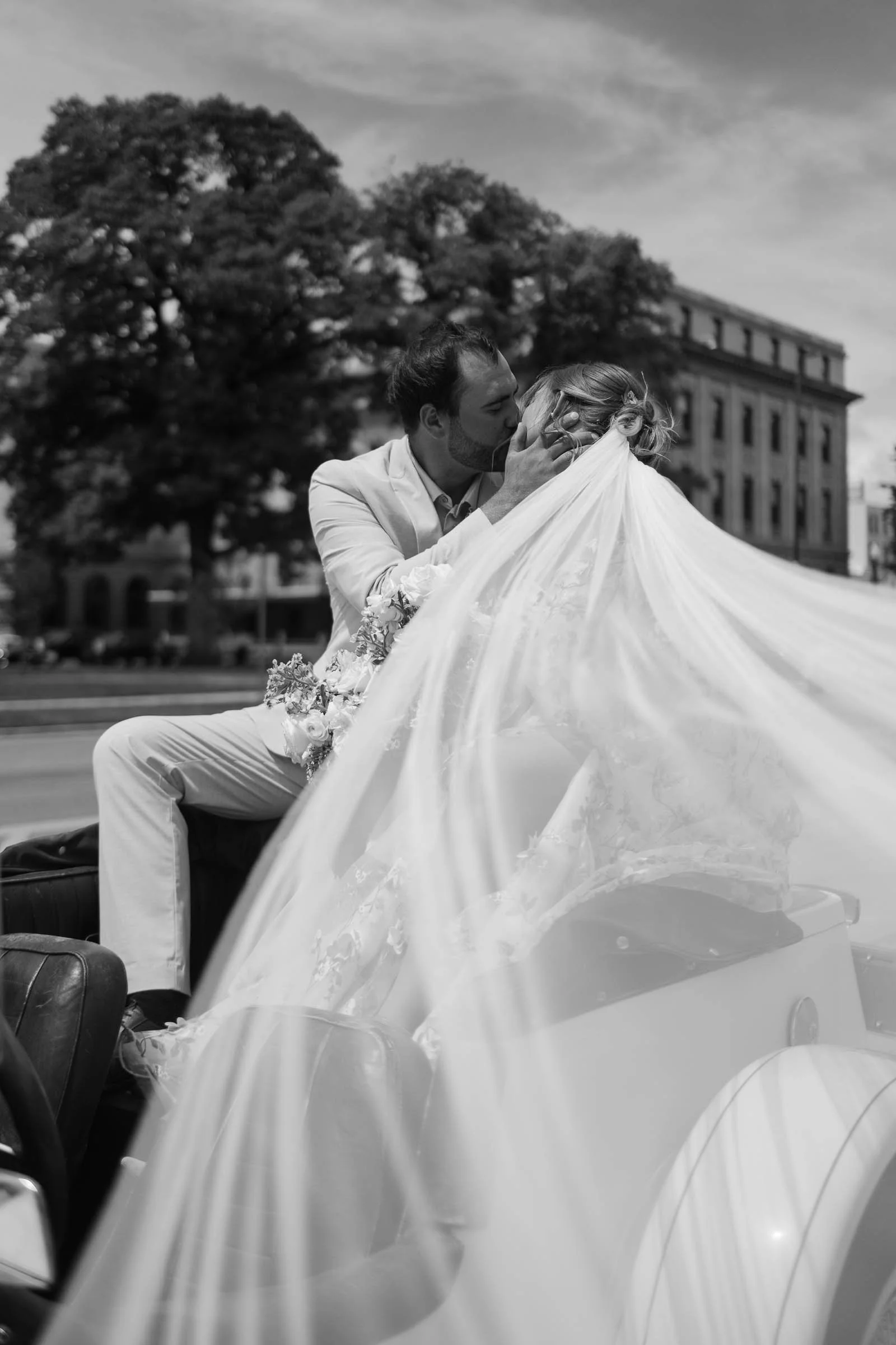  A newlywed couple kisses in the backseat of a convertible as the bride’s veil flows in the wind. The groom wears a light suit, and historic buildings and trees frame the scene. 
