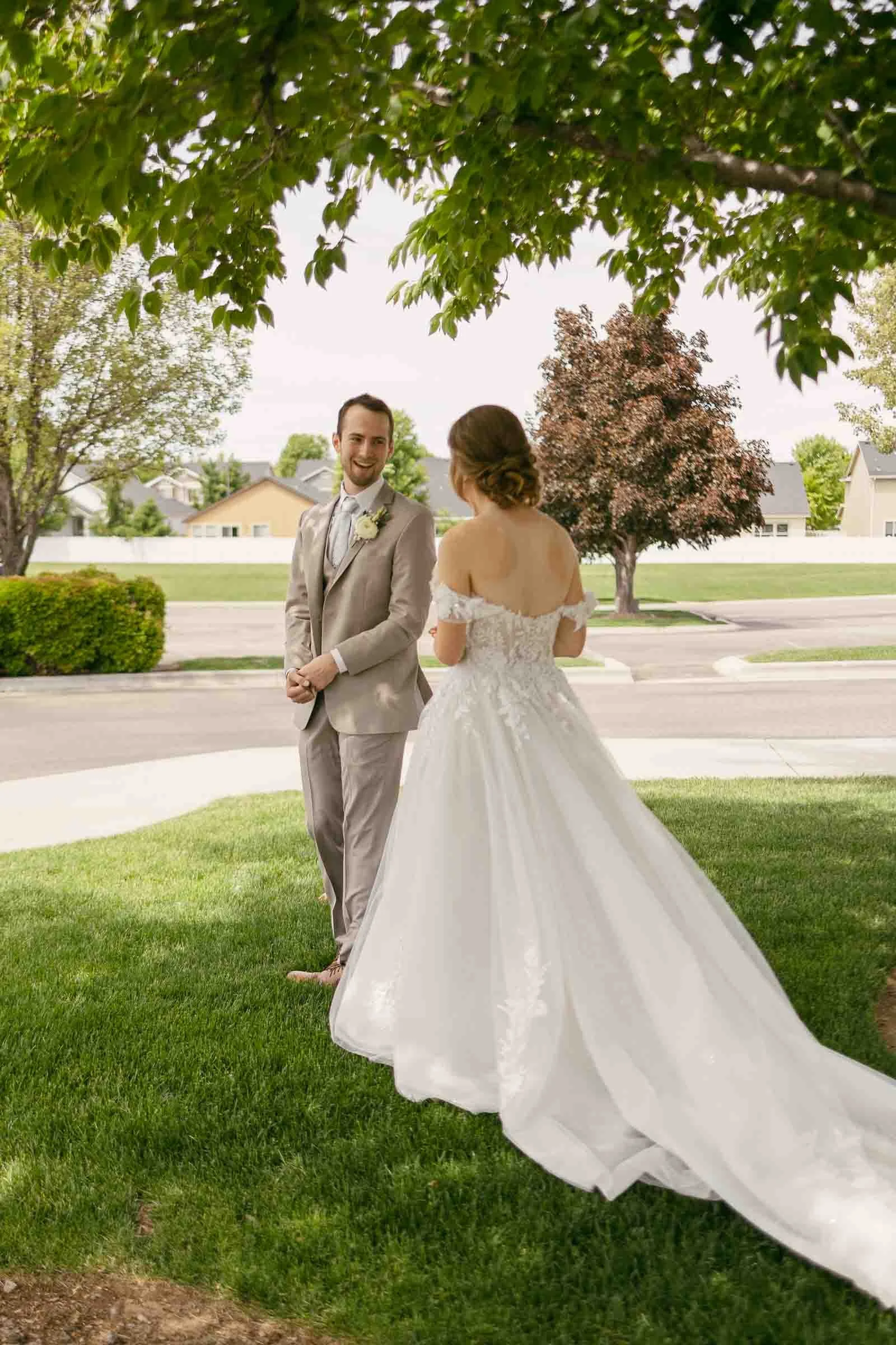 A bride in an off-the-shoulder white gown stands facing a groom in a light brown suit with a white boutonniere. He has just turned around and seen her for the first time. His face is full of wonder and joy. They are outdoors under a tree with green