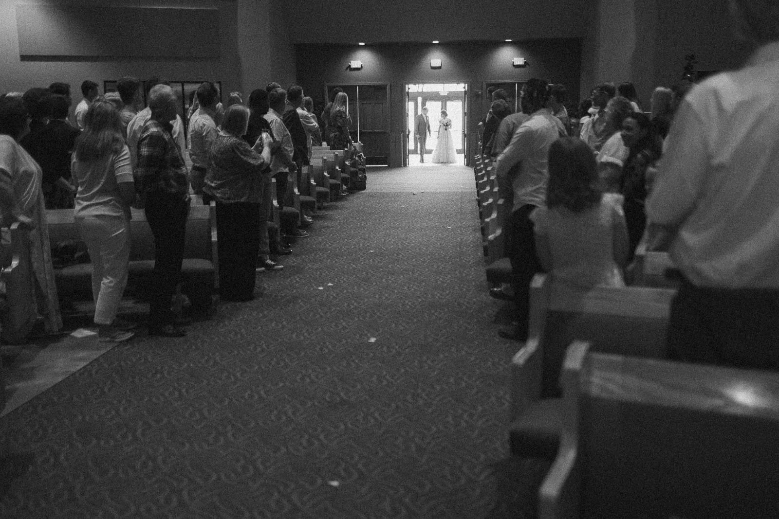 Black and white photo of a wedding ceremony with guests standing on either side of a church aisle. At the end of the aisle, the bride and her dad, are centered in the doorway, illuminated by light from outside.