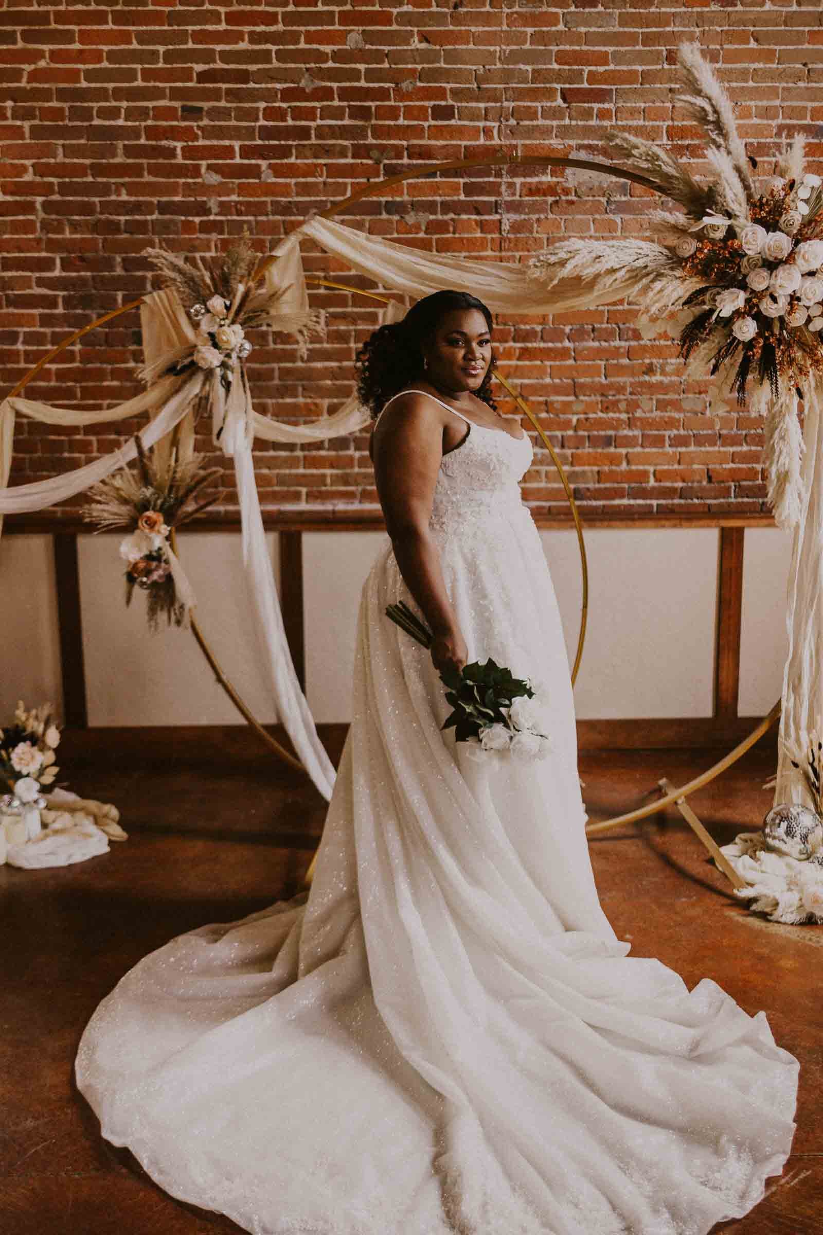  A bride stands confidently in a strapless white wedding dress with a long train, holding a bouquet of white flowers. She poses in front of a rustic brick wall adorned with a circular floral arrangement and decorative dried grasses. 