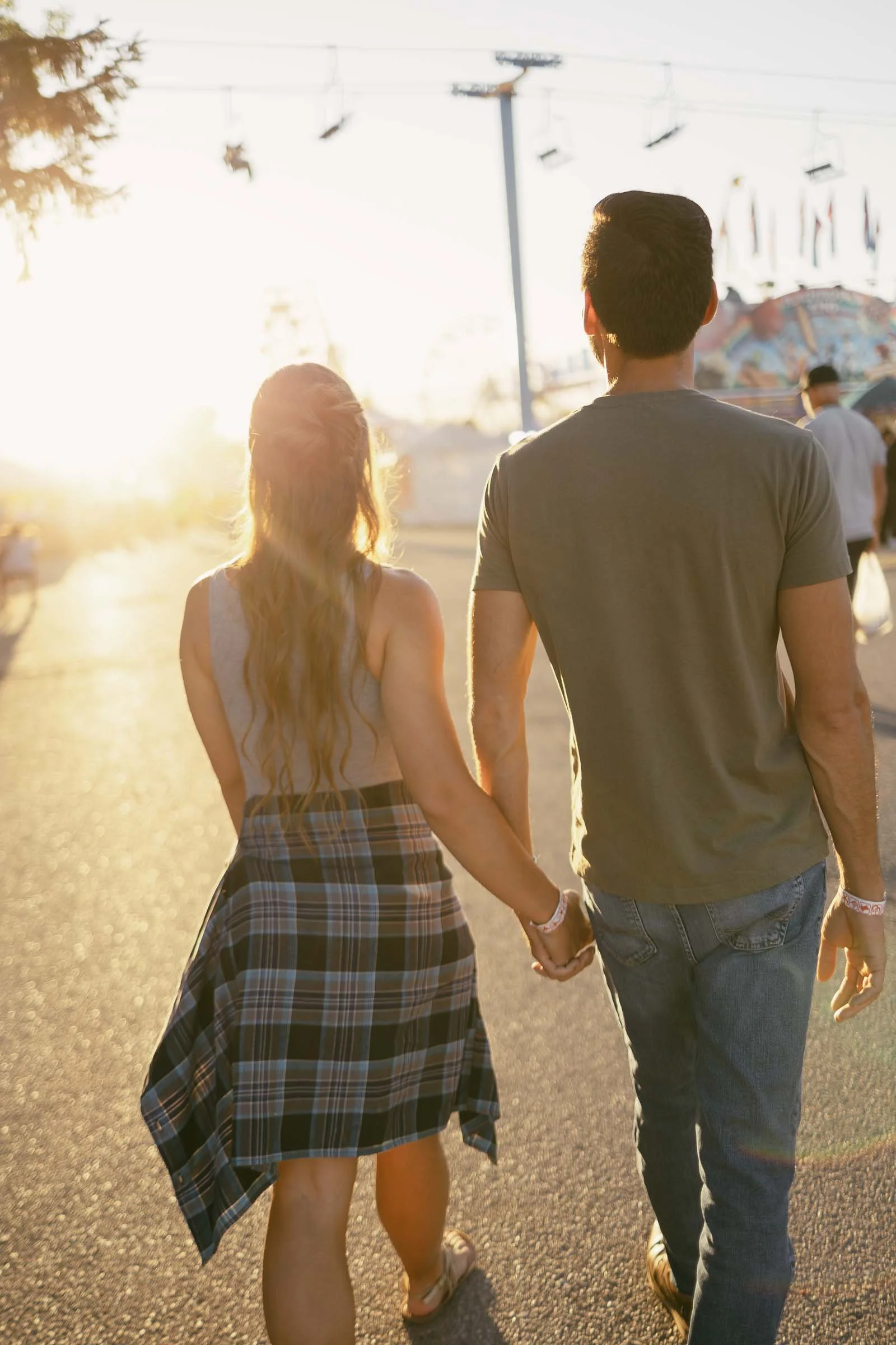 A couple is holding hands while walking on a sunlit path. The woman wears a plaid shirt tied around her waist, and the man is in jeans and a t-shirt. A carnival with rides and flags is visible in the background.