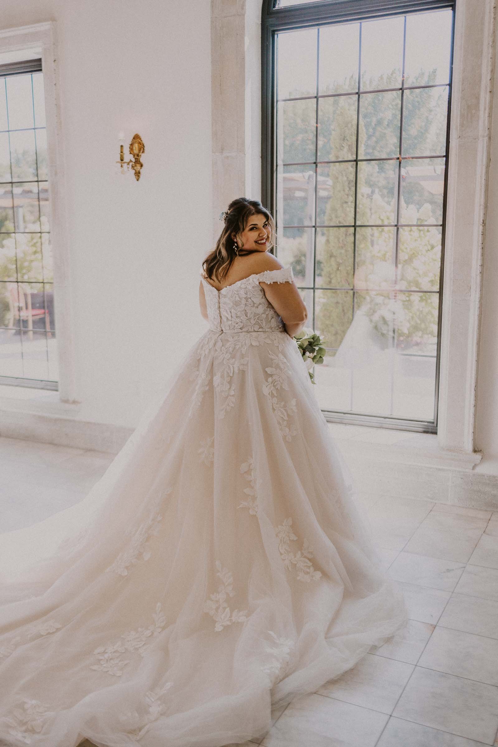  A bride in a flowing white gown with floral details stands near large windows, looking over her shoulder. Sunlight illuminates the room, highlighting the intricate lacework of the dress. The scene is serene and elegant. 