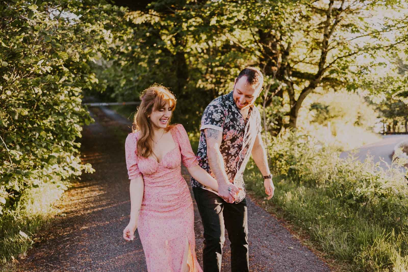 A couple holding hands and walking on a sunlit path surrounded by green trees. The woman is wearing a pink dress, and the man is wearing a patterned shirt and dark pants. They are smiling and looking at each other.