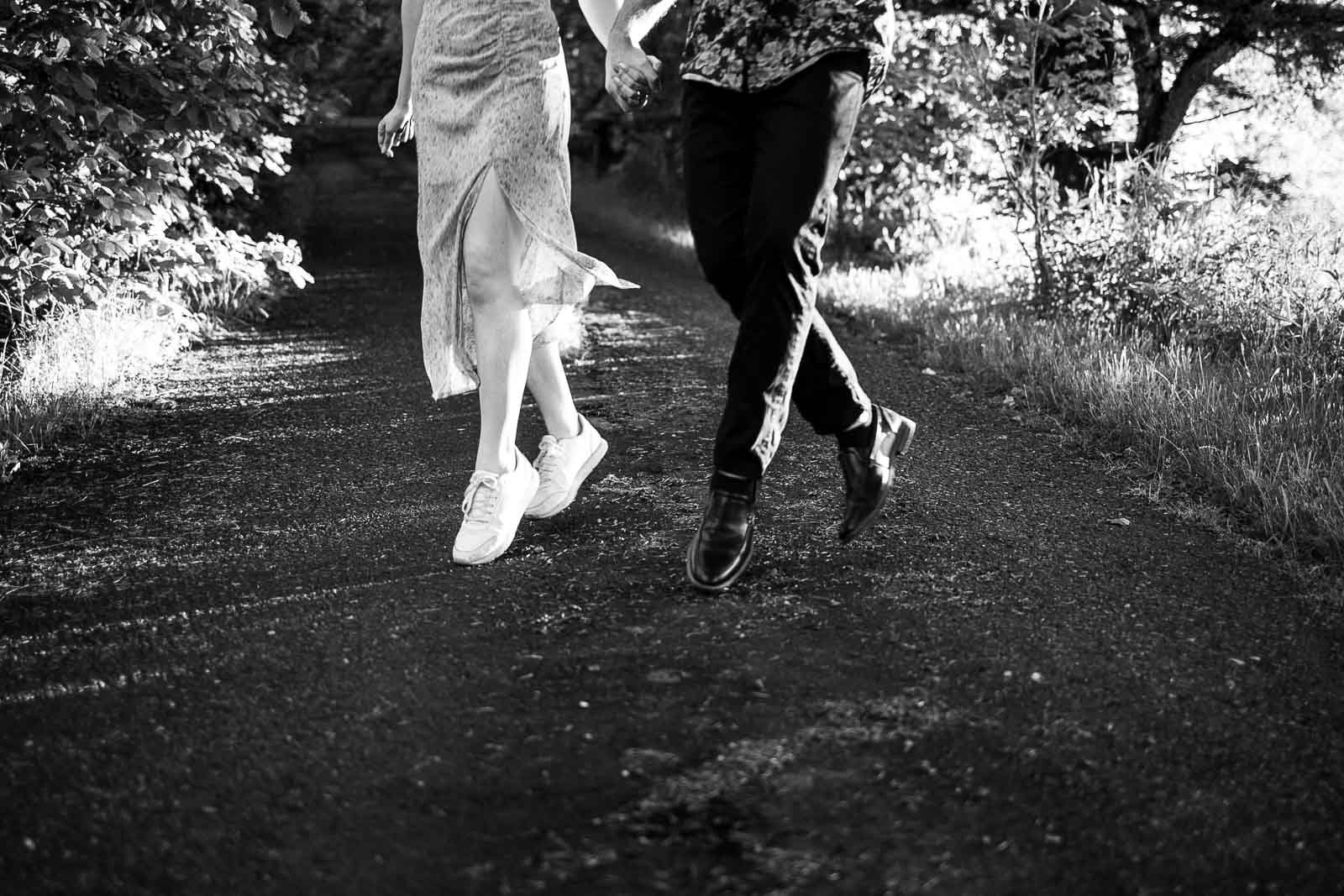 Black and white photo of two people walking hand in hand on a sunlit path. Only their legs are visible. One wears a dress and sneakers, the other wears pants and shoes. Trees line the path.