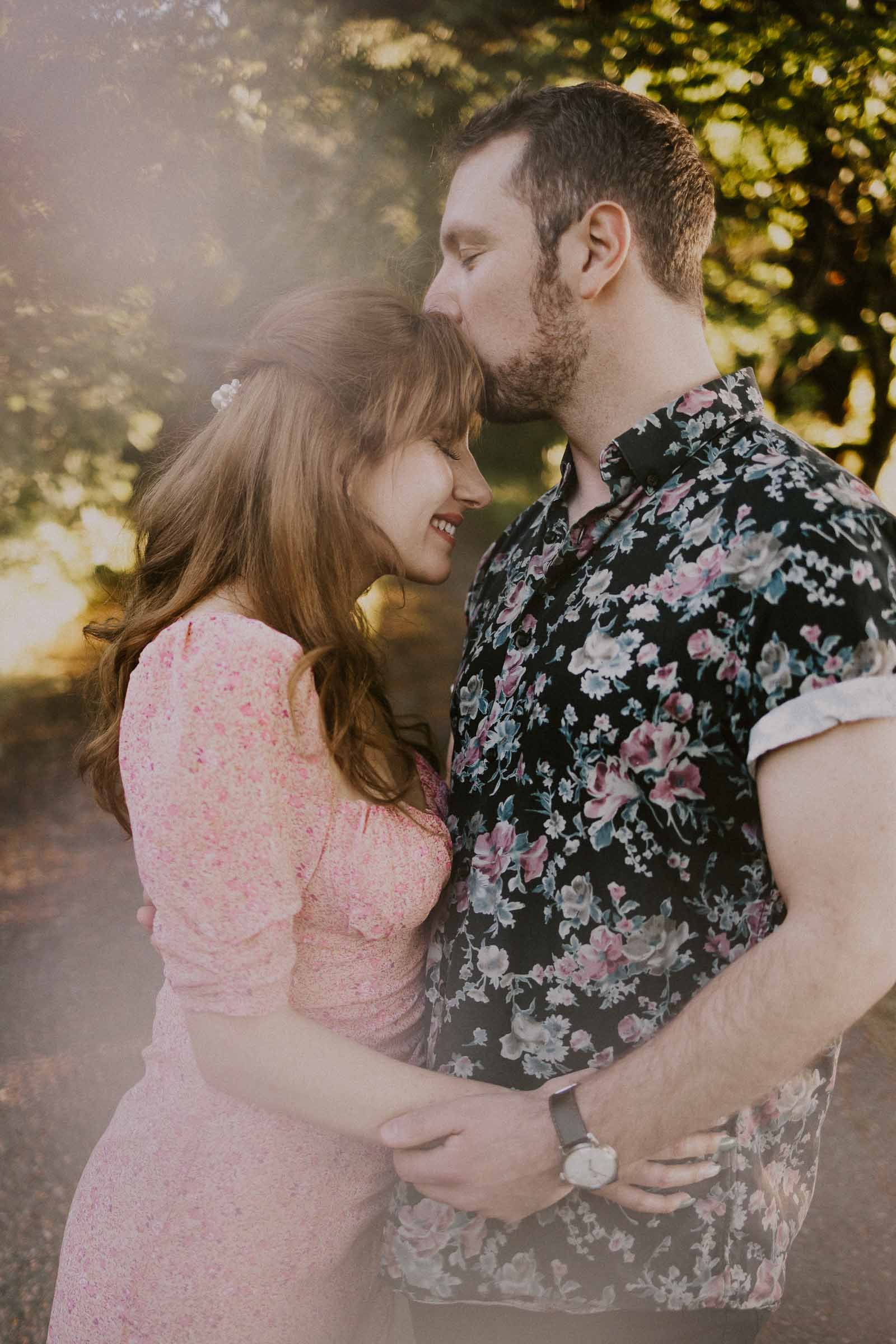 A couple embraces outdoors. The man, in a floral shirt, gently kisses the forehead of the woman, who is wearing a pink dress. They both appear happy and content, surrounded by trees and dappled sunlight.