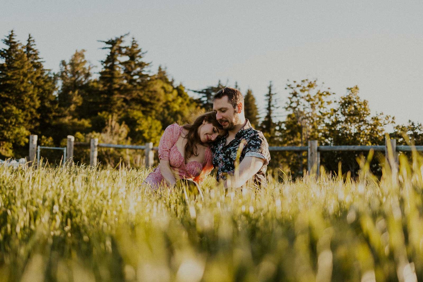 A couple sits together in a grassy field, surrounded by tall grass and trees in the background. The woman leans her head on the man's shoulder, and they appear content. It's a sunny day, casting warm light over the scene.