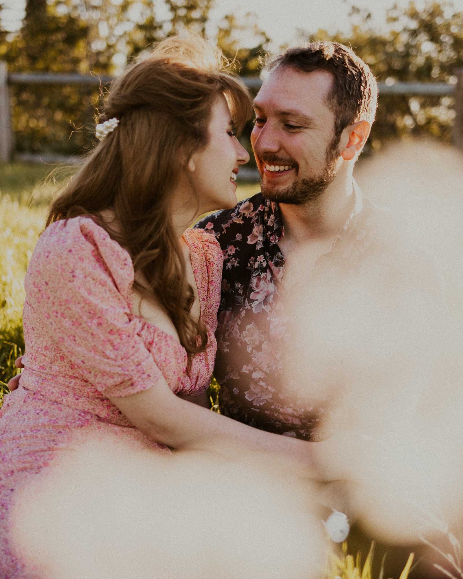 A couple sits closely together on grass, smiling warmly at each other. The woman wears a pink dress, and the man has a floral shirt. Soft sunlight and blurred foreground elements create a dreamy, romantic atmosphere.