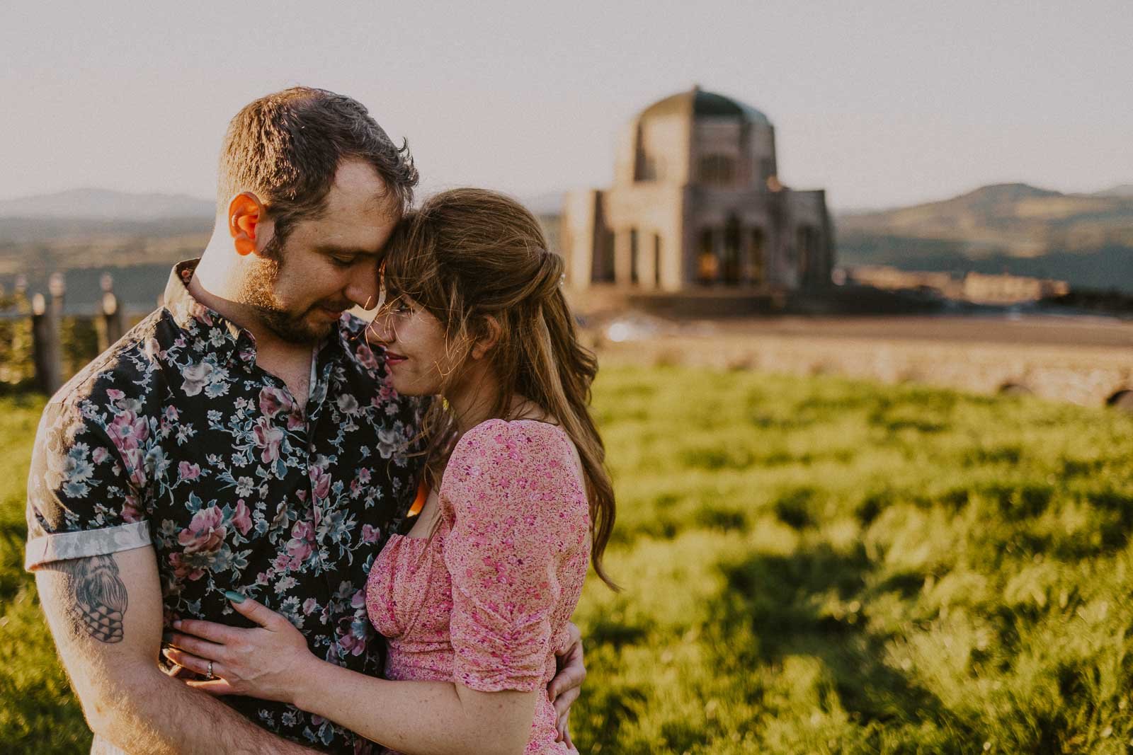 A couple embraces in a grassy field, with a historical domed stone structure in the background. The man wears a floral shirt, and the woman wears a pink dress. The setting is bathed in warm, golden light.