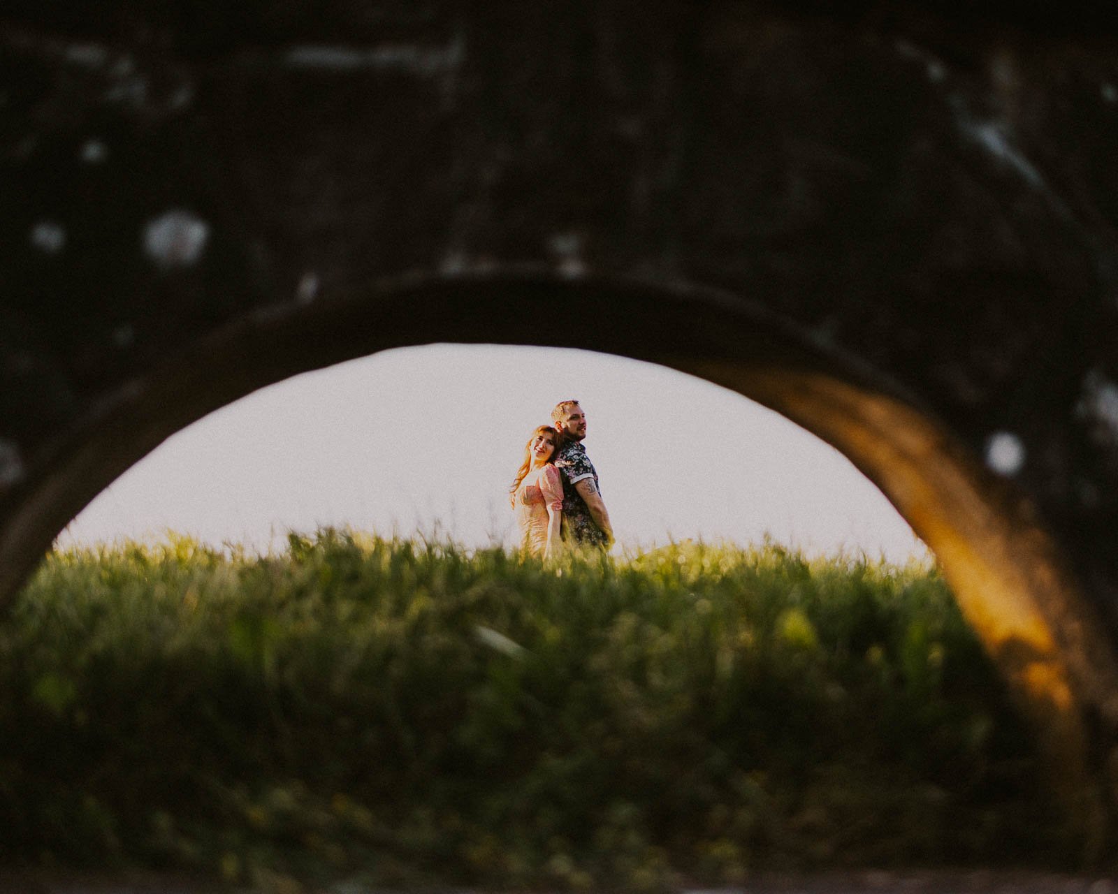 A couple stands closely together in a field of tall grass. They are visible through a large circular opening in a stone structure, with soft lighting enhancing the serene scene.