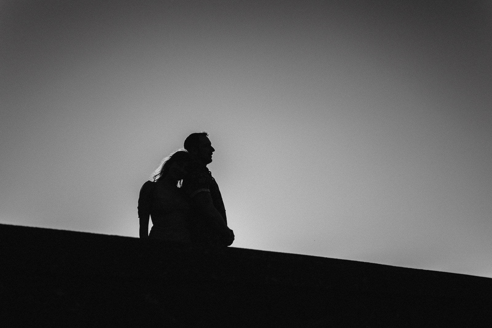 Silhouetted image of a couple standing closely together, with the woman resting her head on the man's shoulder. They are on an elevated surface against a clear, bright sky, creating a dramatic contrast between their outlines and the background.