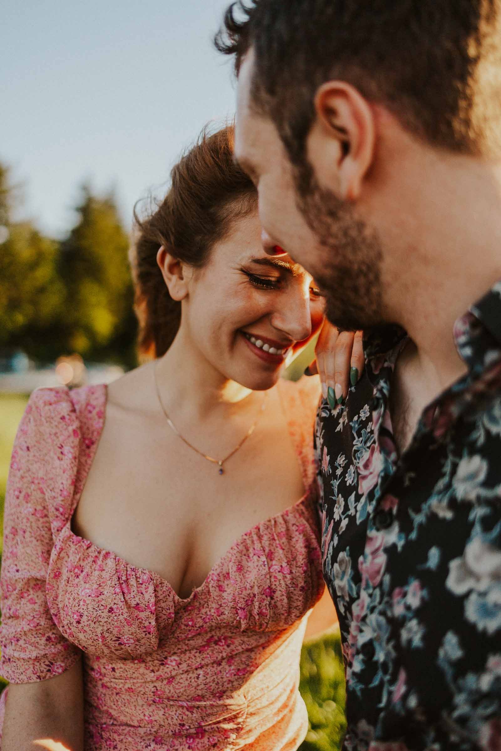 A couple stands close together in a sunny outdoor setting, gazing at each other and smiling. The woman wears a pink floral dress, and the man wears a dark floral shirt. Green grass and blurred trees are in the background.