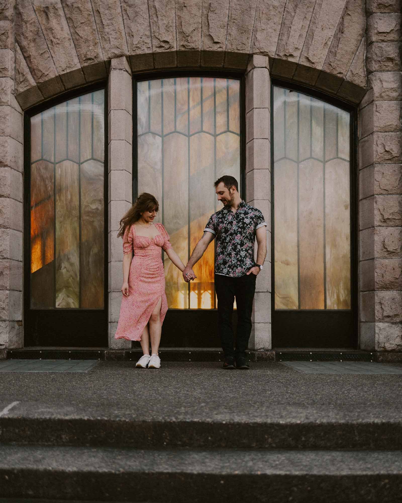 A couple holding hands stands on stone steps in front of a building with large arched windows. The woman wears a pink dress, and the man is in a patterned shirt and black pants. The windows reflect a warm, glowing light.