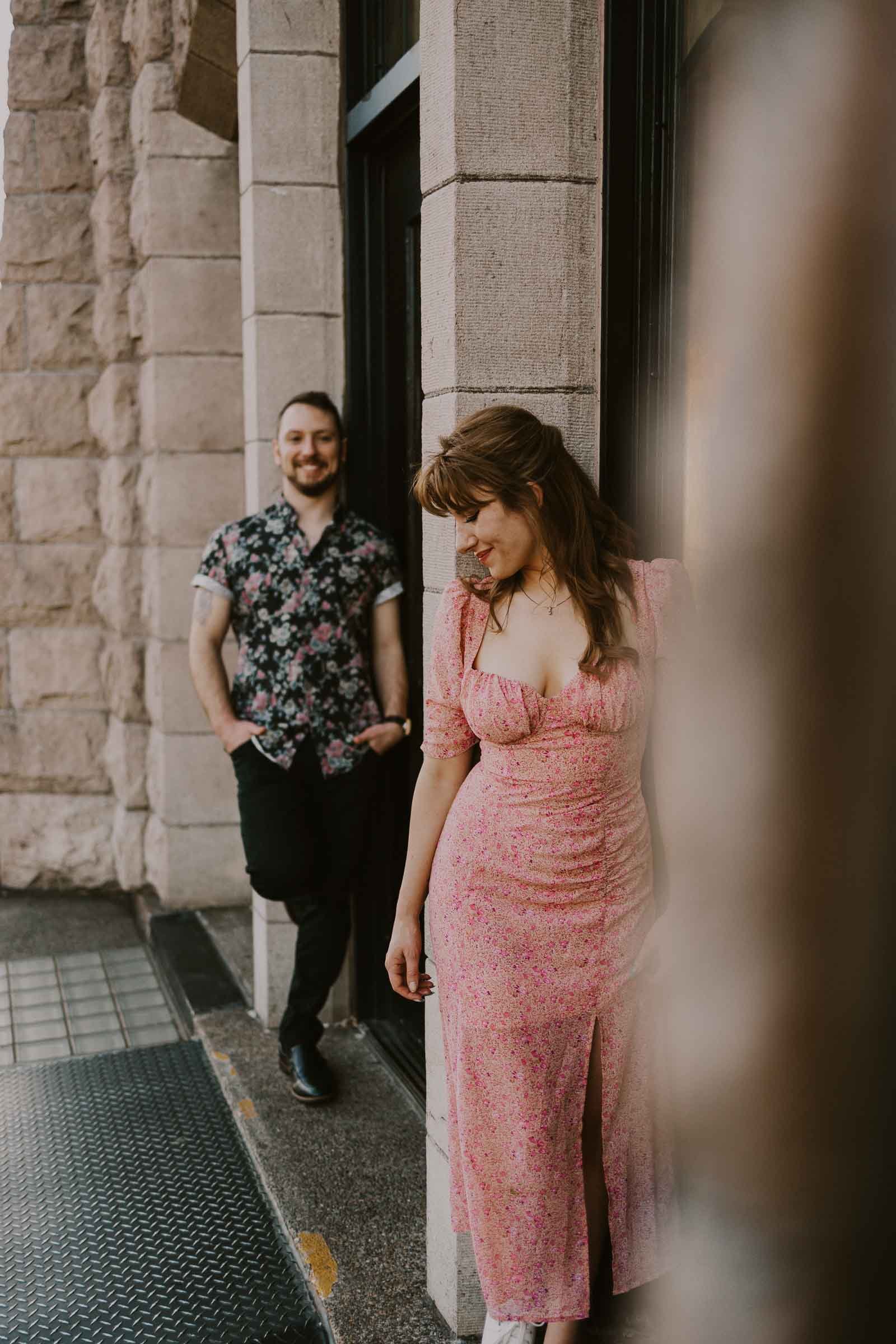 A woman in a pink floral dress leans against a stone building, smiling and looking down. In the background, a man in a black floral shirt stands casually, smiling. The setting appears to be an urban area with textured walls.