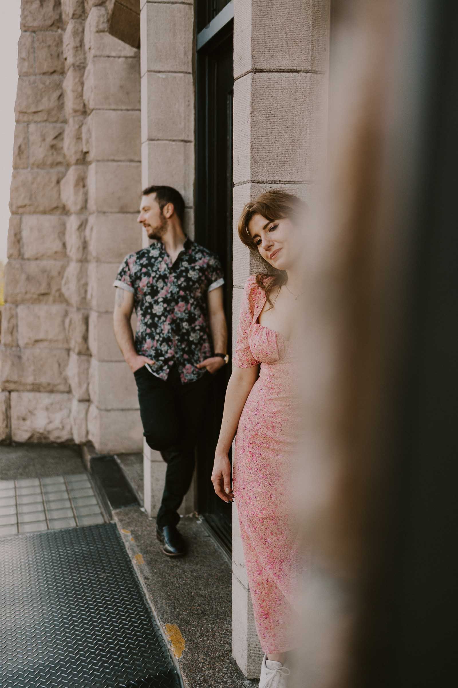 A woman in a pink floral dress leans against a building, looking downward, while a man in a floral shirt stands nearby with his hands in his pockets, gazing into the distance. Stone walls frame the scene.