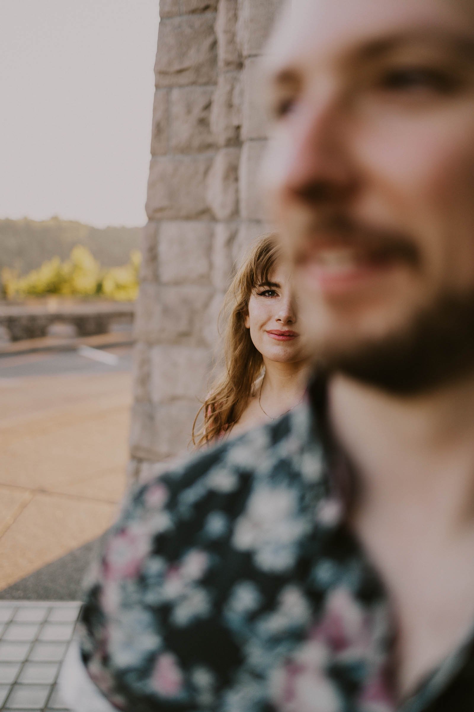 A woman smiles while standing behind a man in focus, near a stone wall. The background features a road and greenery. The man’s floral shirt is prominent in the foreground, slightly blurred.