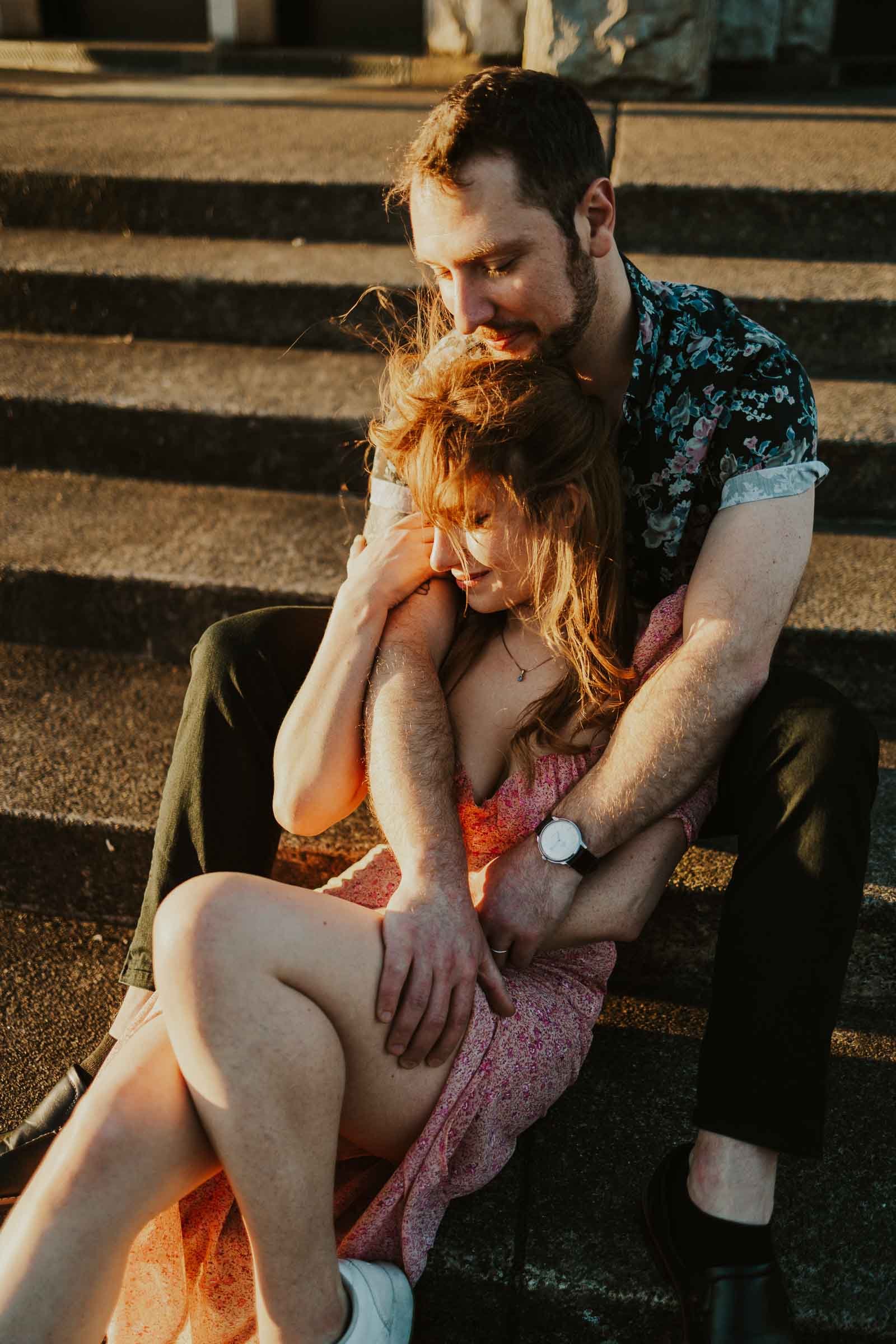 A couple sits closely on outdoor steps, embracing each other. The woman, wearing a pink dress, leans into the man's arms. The man is wearing a patterned shirt and watches the sunset. The warm light creates a cozy atmosphere.