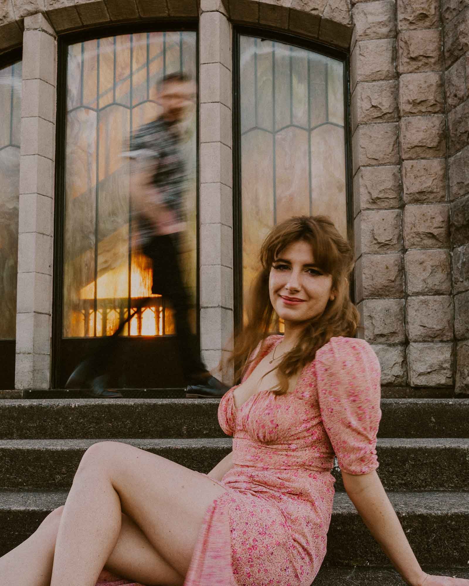 A woman in a pink dress sits on stone steps, smiling at the camera. Behind her, a blurred man walks past large arched windows with warm lighting. The building has stone walls, creating a historic atmosphere.