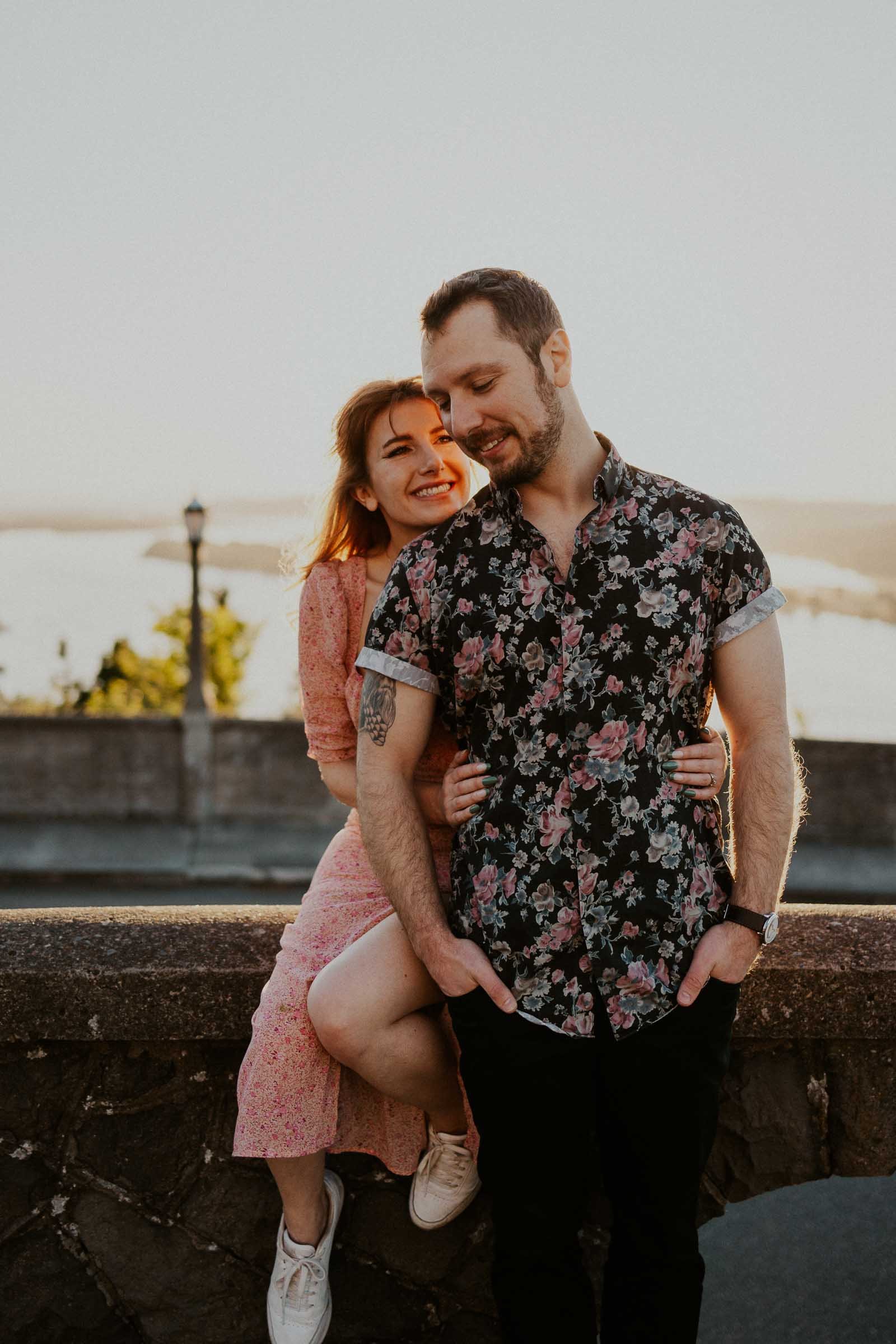 A woman in a pink dress sits on a ledge, smiling and embracing a man standing next to her. He wears a floral shirt and has his hands in his pockets. They are outdoors during sunset, with a blurred background of water and trees.