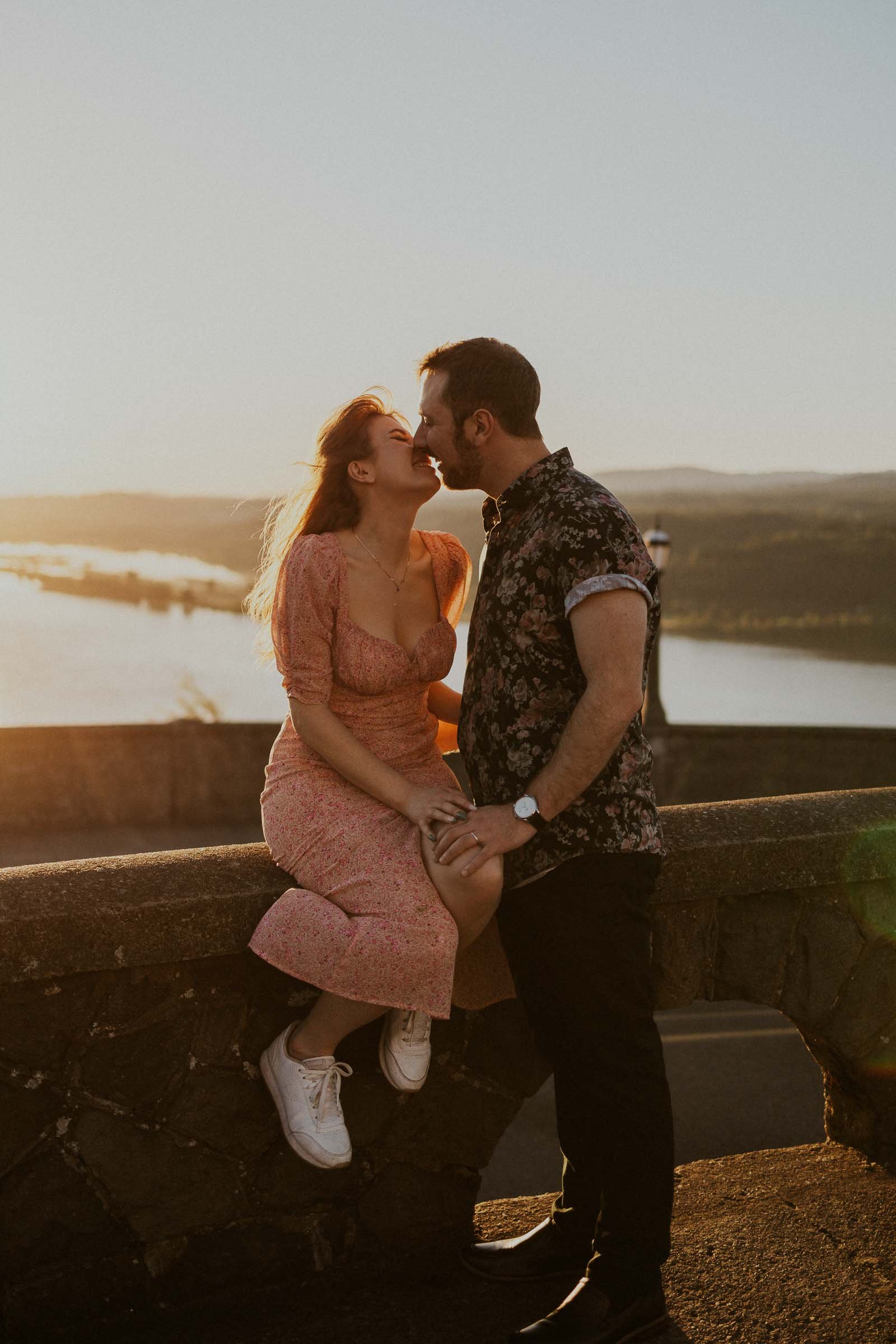 A couple is sharing a tender moment at sunset on a stone wall overlooking a scenic river. The woman in a floral dress is sitting while the man in a patterned shirt stands, with warm sunlight illuminating them from behind.