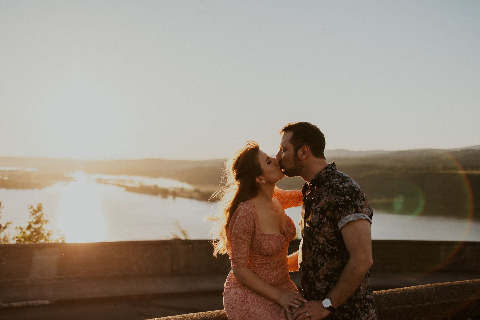 A couple kisses while standing on a ledge overlooking a vast landscape with a river. The sun sets in the background, casting a warm, golden light over the scene. The woman wears a pink dress, and the man is in a floral shirt.