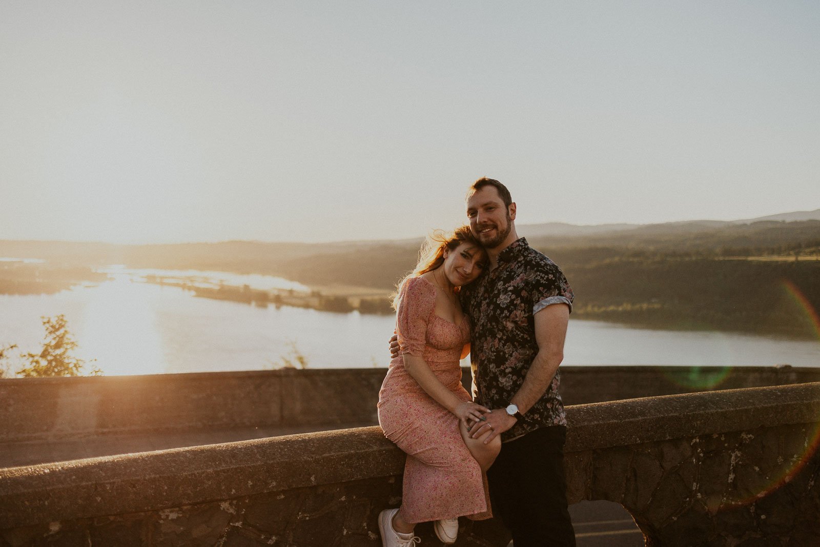 A couple embraces while sitting on a stone wall, with a scenic view of a river and hills in the background during sunset. The woman leans her head on the man's shoulder, both smiling. The sunlight casts a warm glow over the scene.