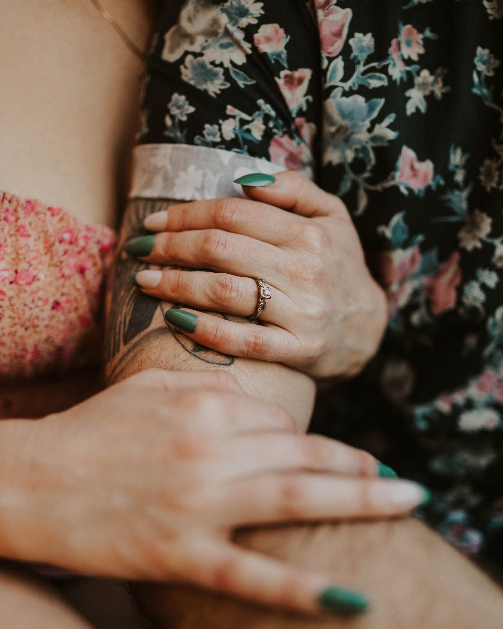 A close-up of two people embracing. One person has their hand on the other's tattooed arm, showcasing green nail polish and a ring. Both wear floral-patterned clothing, creating a soft and intimate scene.