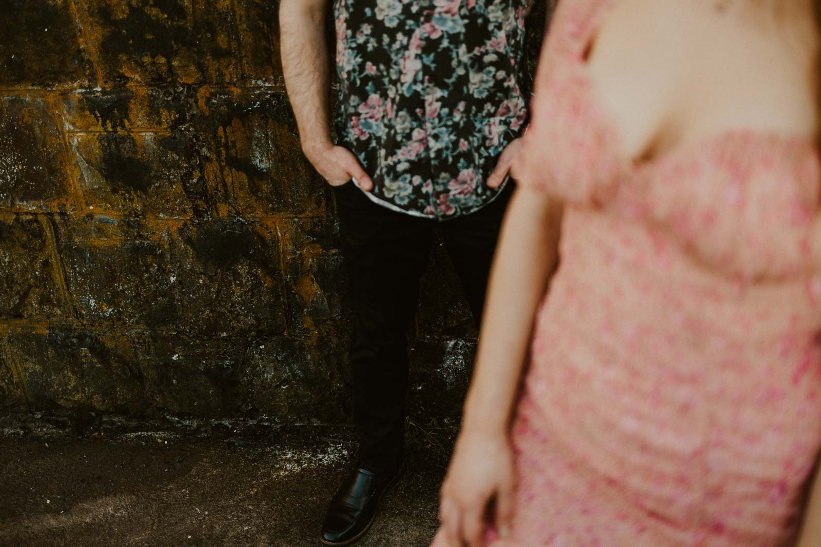 A person in a floral shirt stands against a textured wall, partly visible with hands in pockets. In the foreground, another person in a pink floral dress is out of focus, only partially shown. The scene has a vintage and relaxed vibe.