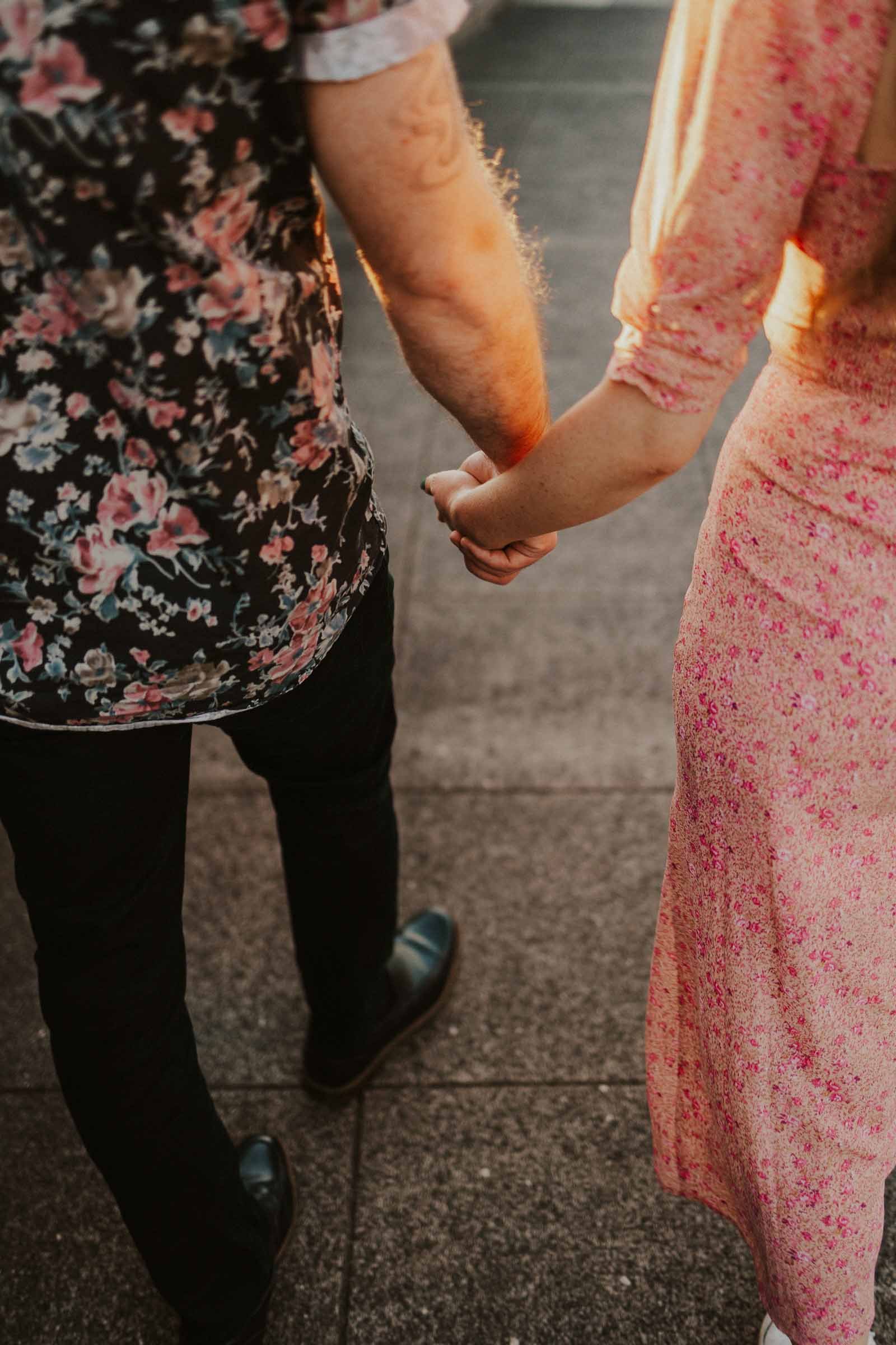 A couple holds hands while walking. The person on the left wears a black floral shirt and dark pants, and the person on the right wears a pink floral dress. They're on a concrete pathway, with sunlight casting shadows.