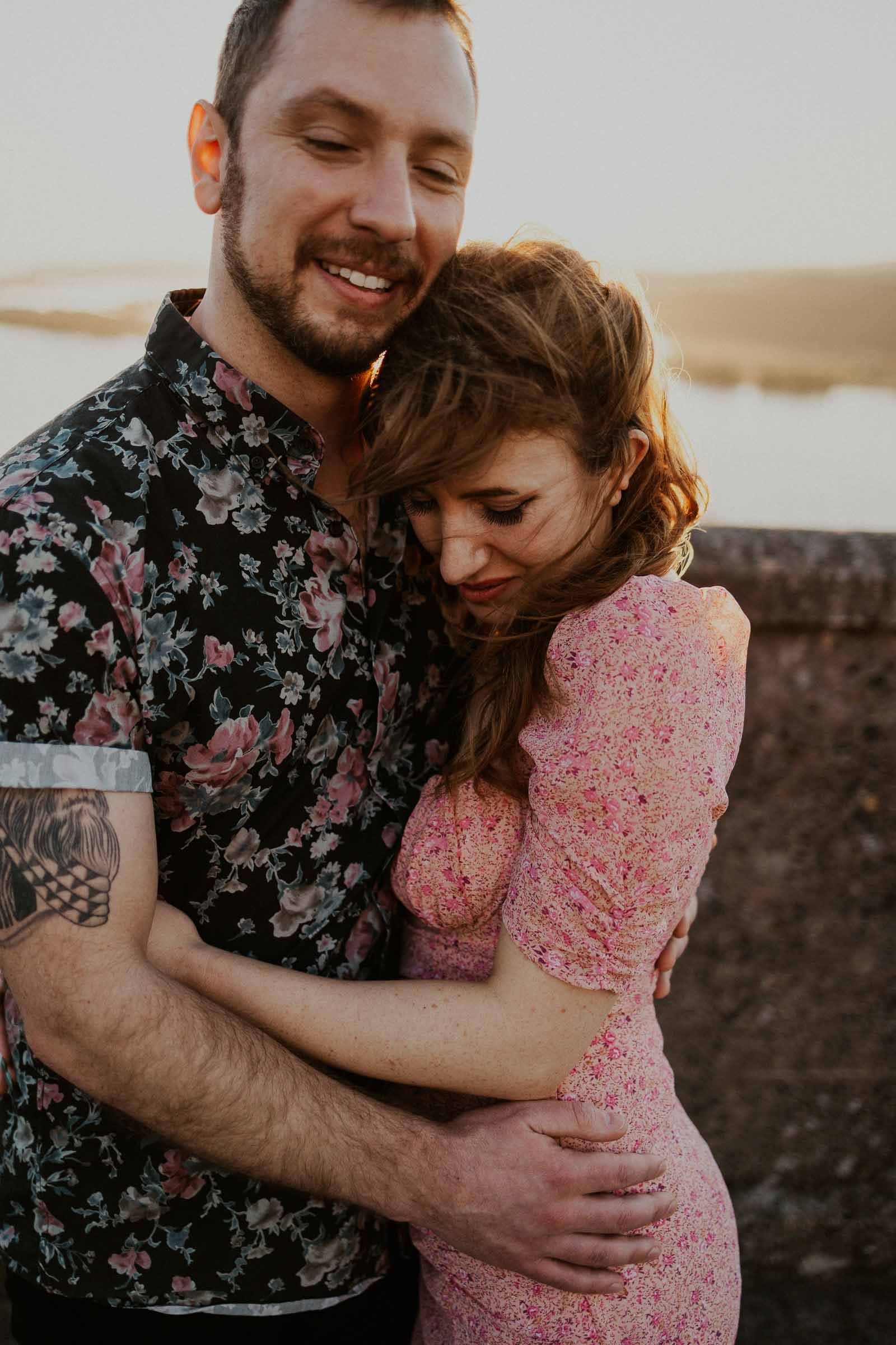 A couple embraces on a windy day by a stone wall, smiling and wearing floral-patterned clothes. The sun sets in the background, casting a warm glow over the scene.