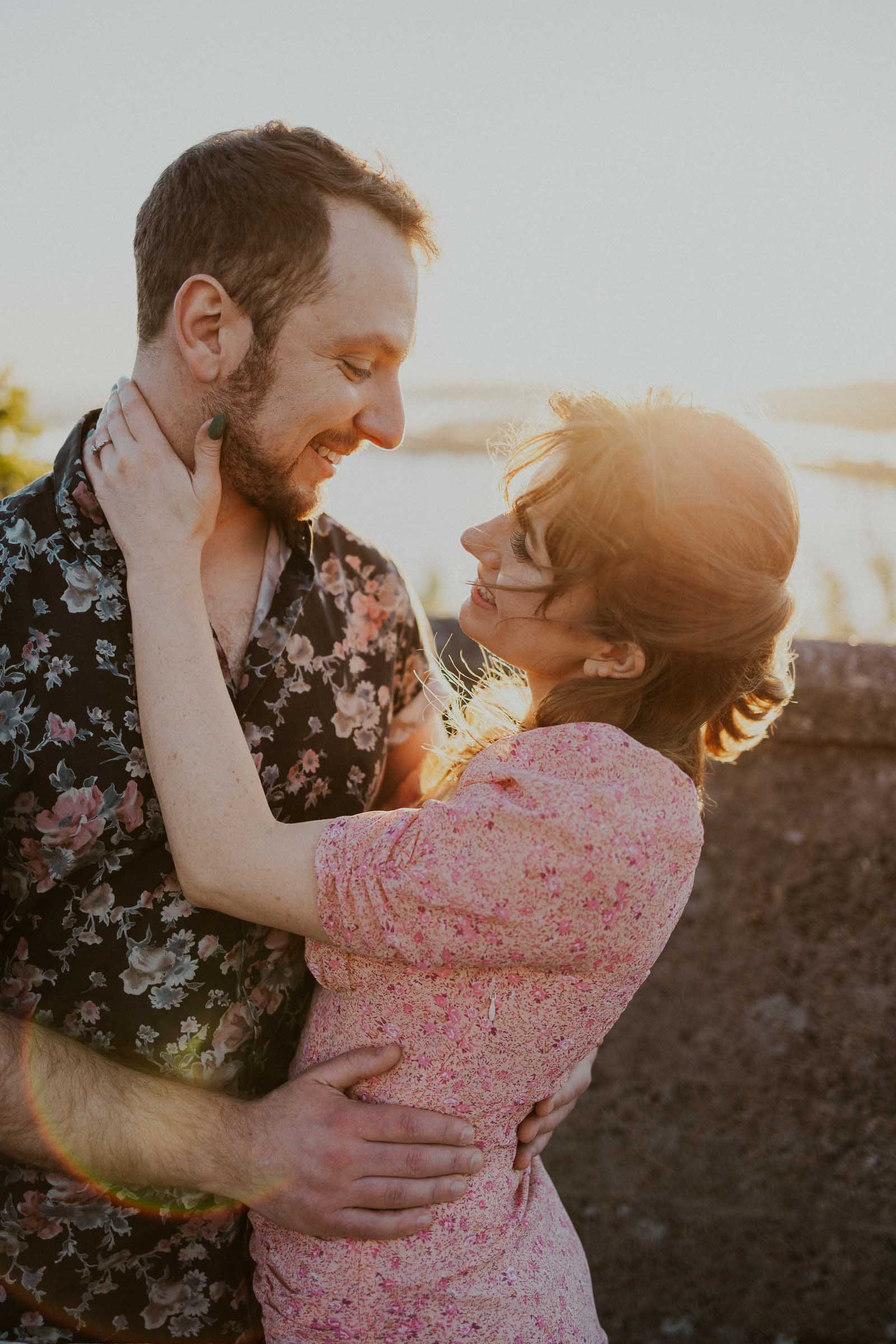 A couple stands close together, smiling at each other. The man wears a floral shirt, while the woman is in a pink dress. The sun sets in the background, casting a warm glow over the scene.
