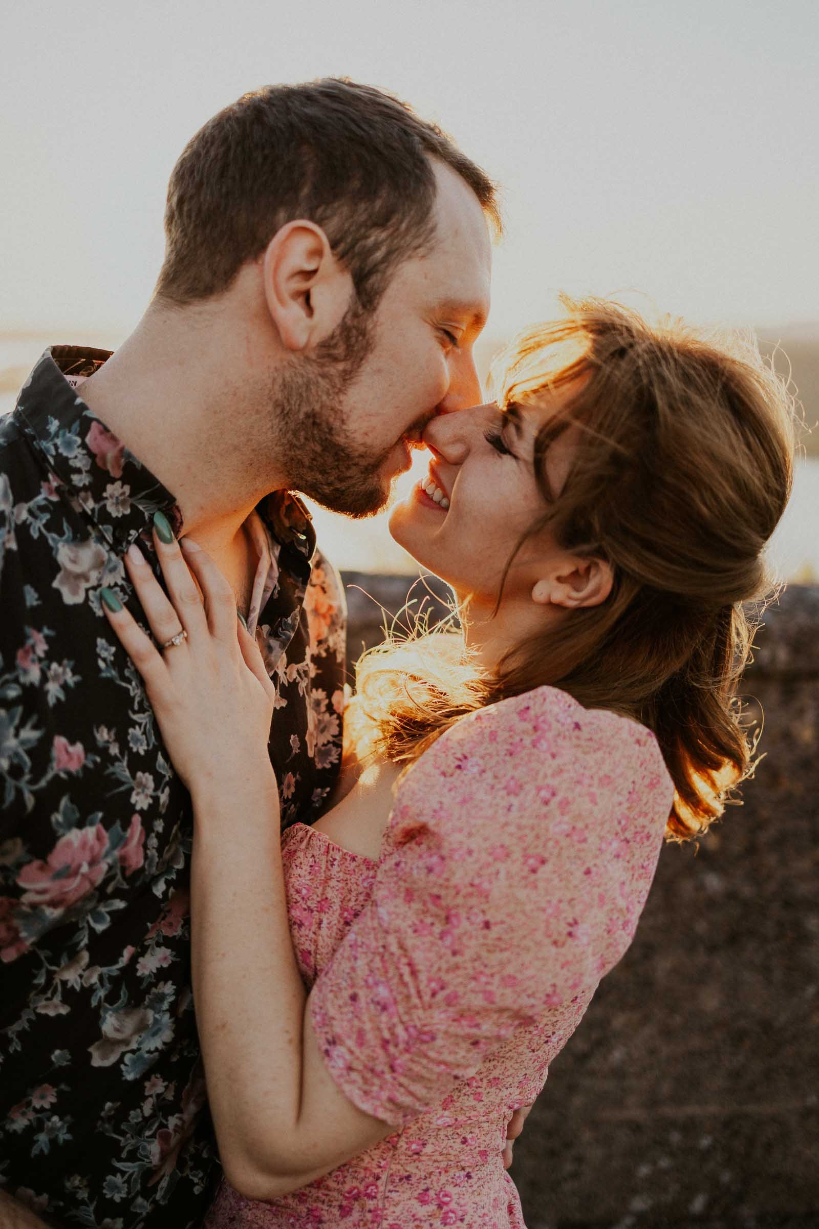 A couple sharing a joyful and intimate moment, standing close and smiling at each other. The woman is wearing a pink floral dress, and the man is in a dark floral shirt. They are outdoors with soft sunlight in the background.