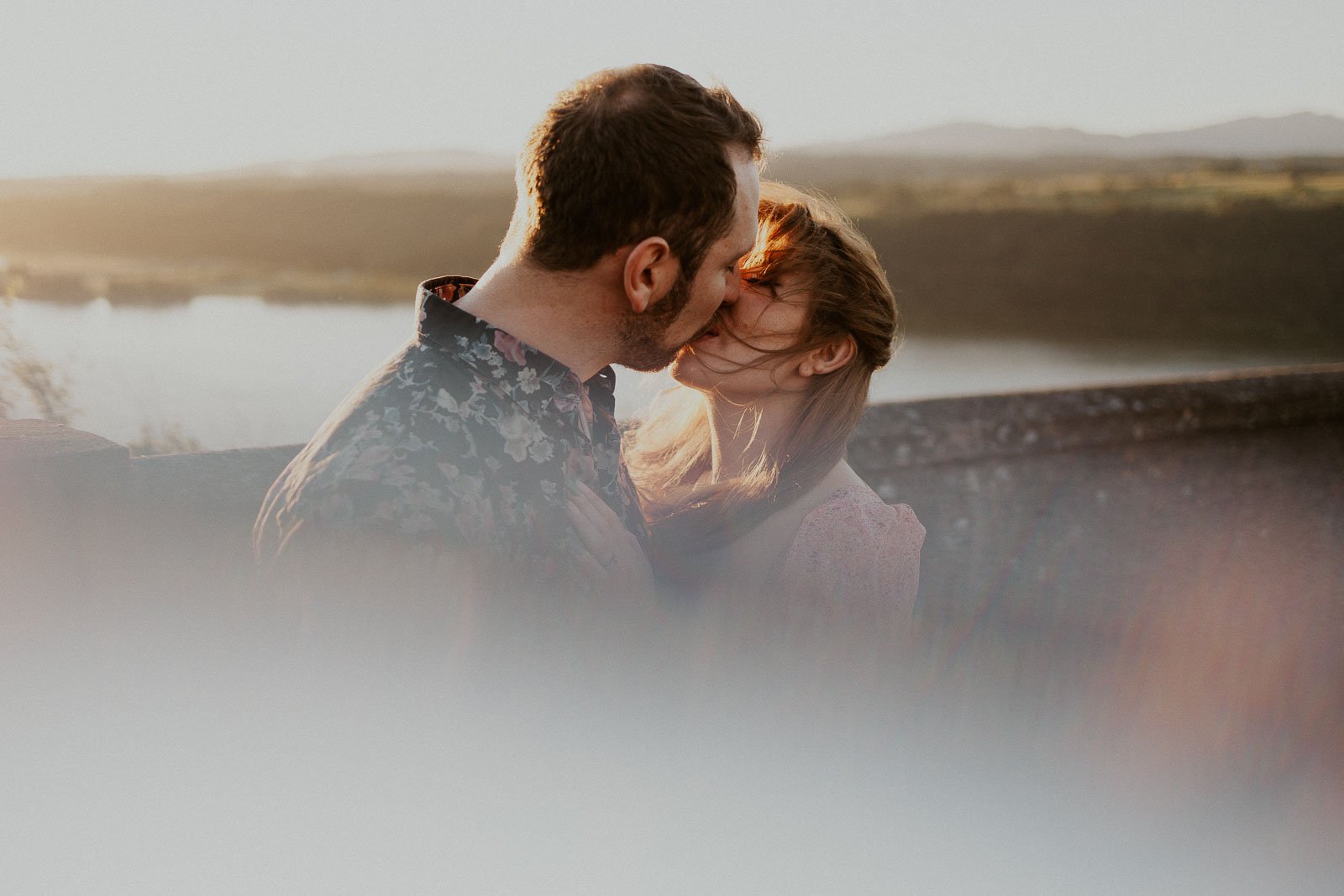 A couple shares a kiss at sunset, standing near a stone wall with a serene lake and hills in the background. The soft sunlight creates a warm, dreamy atmosphere, with gentle lens flare adding to the tranquility of the scene.