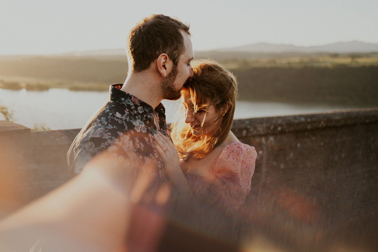 A couple stands on a stone balcony overlooking a serene lake and hills at sunset. The man kisses the woman's forehead as they embrace, both illuminated by the golden sunlight. Soft focus and a lens flare add warmth to the scene.