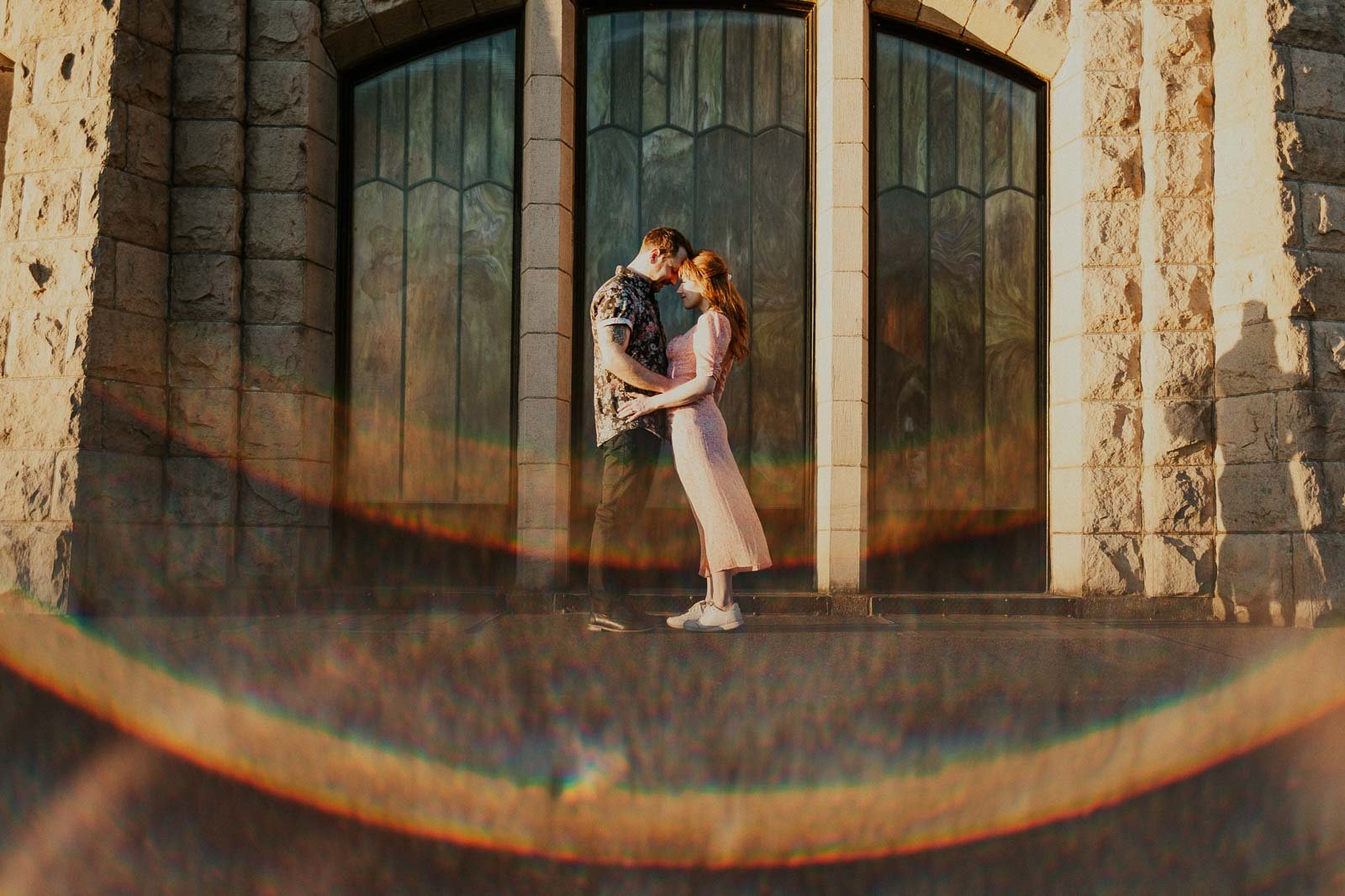 A couple stands close together in front of a grand stone building with large arched windows. They gaze at each other, enveloped in warm sunlight and surrounded by a colorful lens flare. The woman wears a pink dress, and the man wears a patterned shi