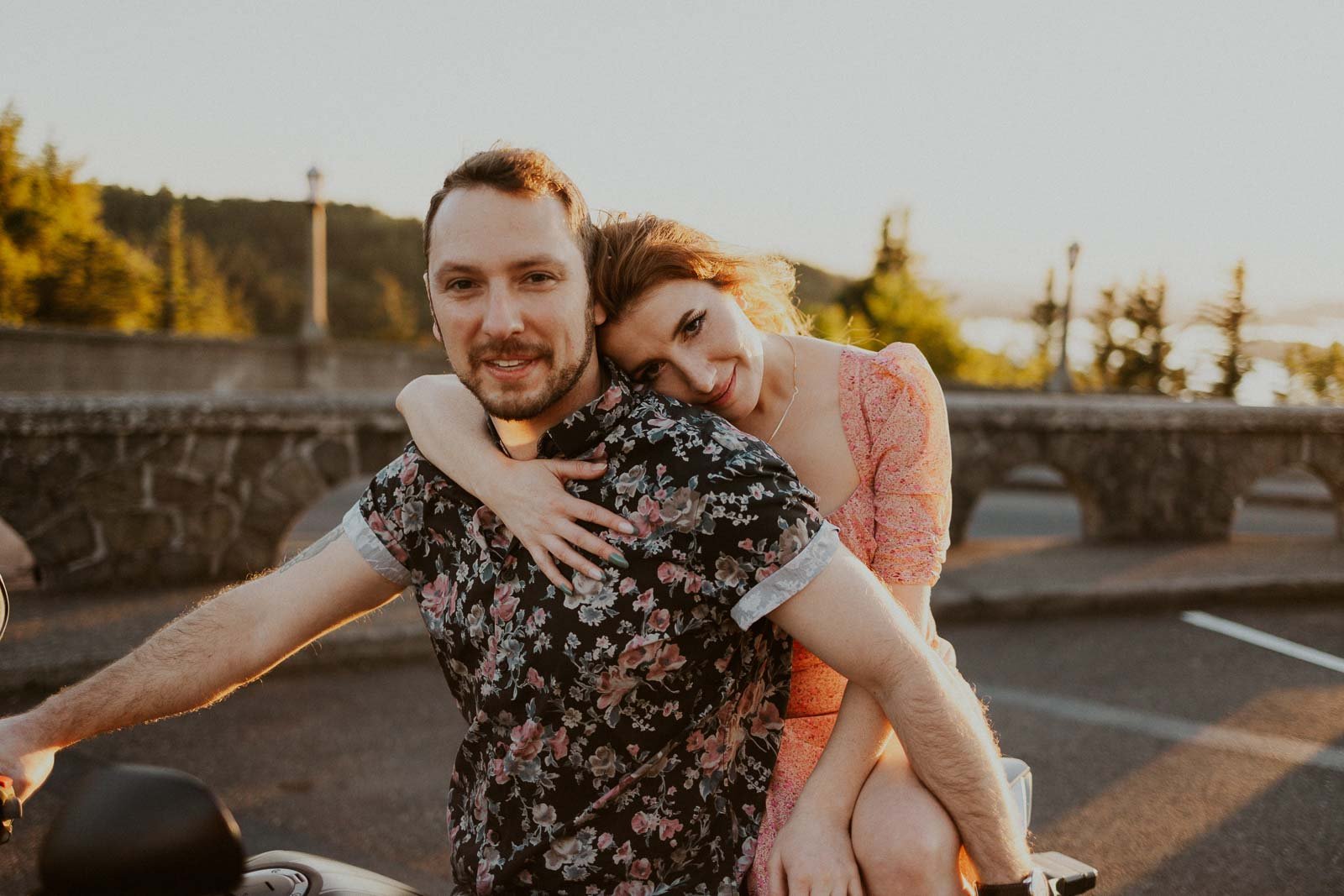 A woman in a pink dress hugs a man in a floral shirt from behind as they sit on a motorcycle. They are outdoors with a sunny backdrop of green trees and a stone wall. The atmosphere is warm and relaxed.