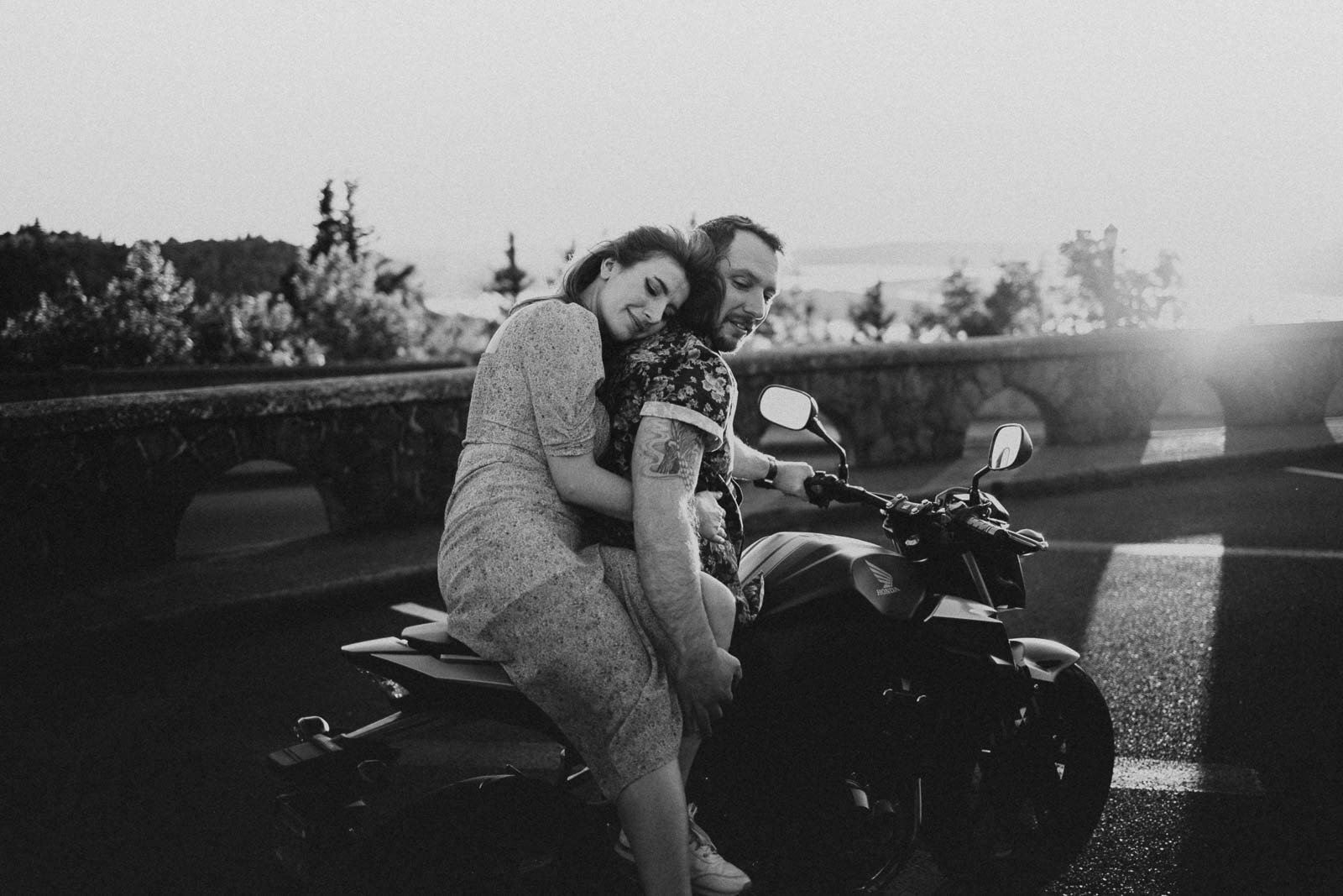 A black-and-white photo of a couple sitting on a motorcycle, smiling and embracing. The background shows a stone wall and trees with a distant view of water and sunset.