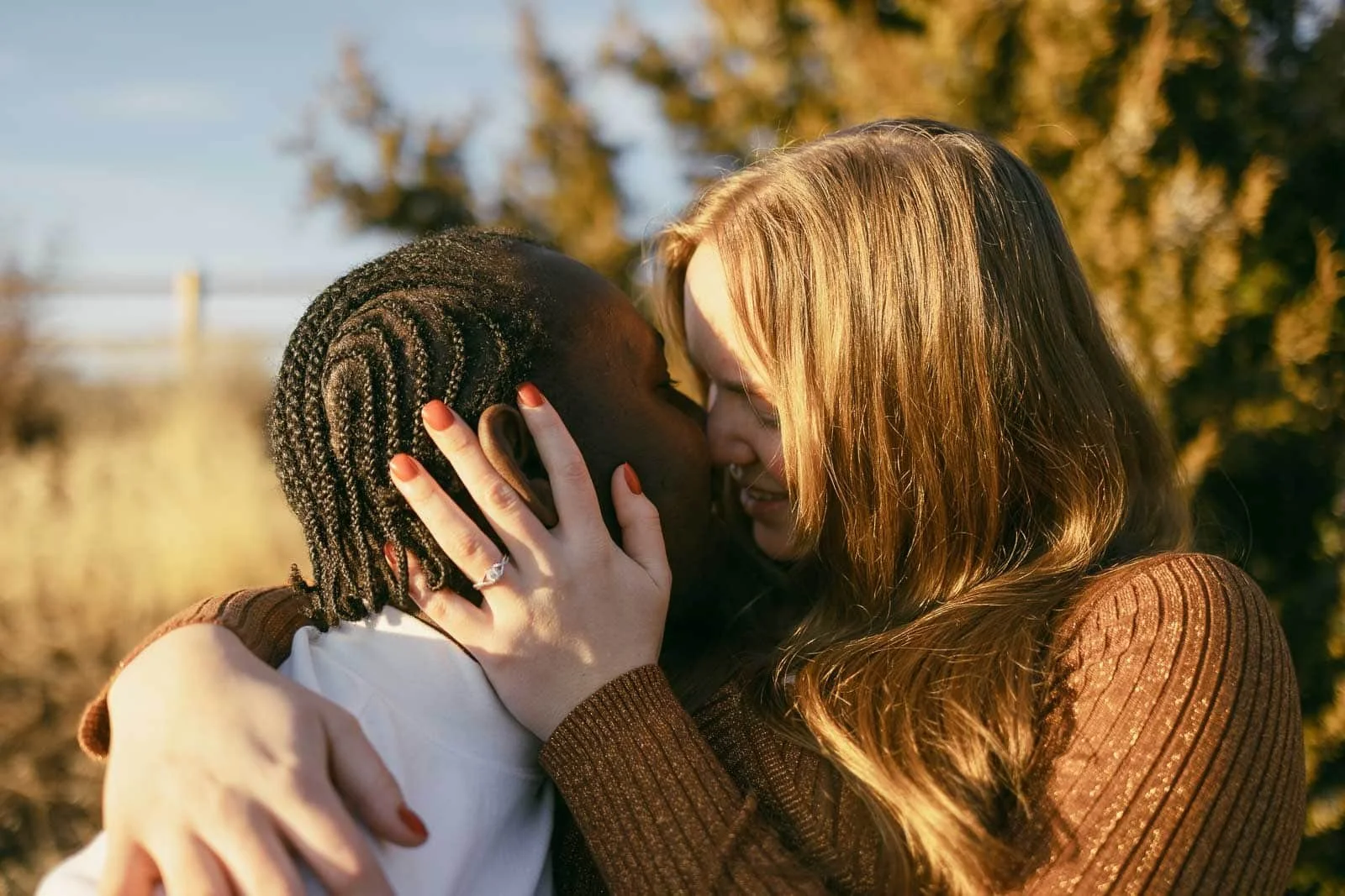  Two people embracing closely outdoors, the woman is going in for a kiss. One has braided hair and the other has long, light brown hair. They are smiling and touching foreheads, surrounded by sunlit greenery. 