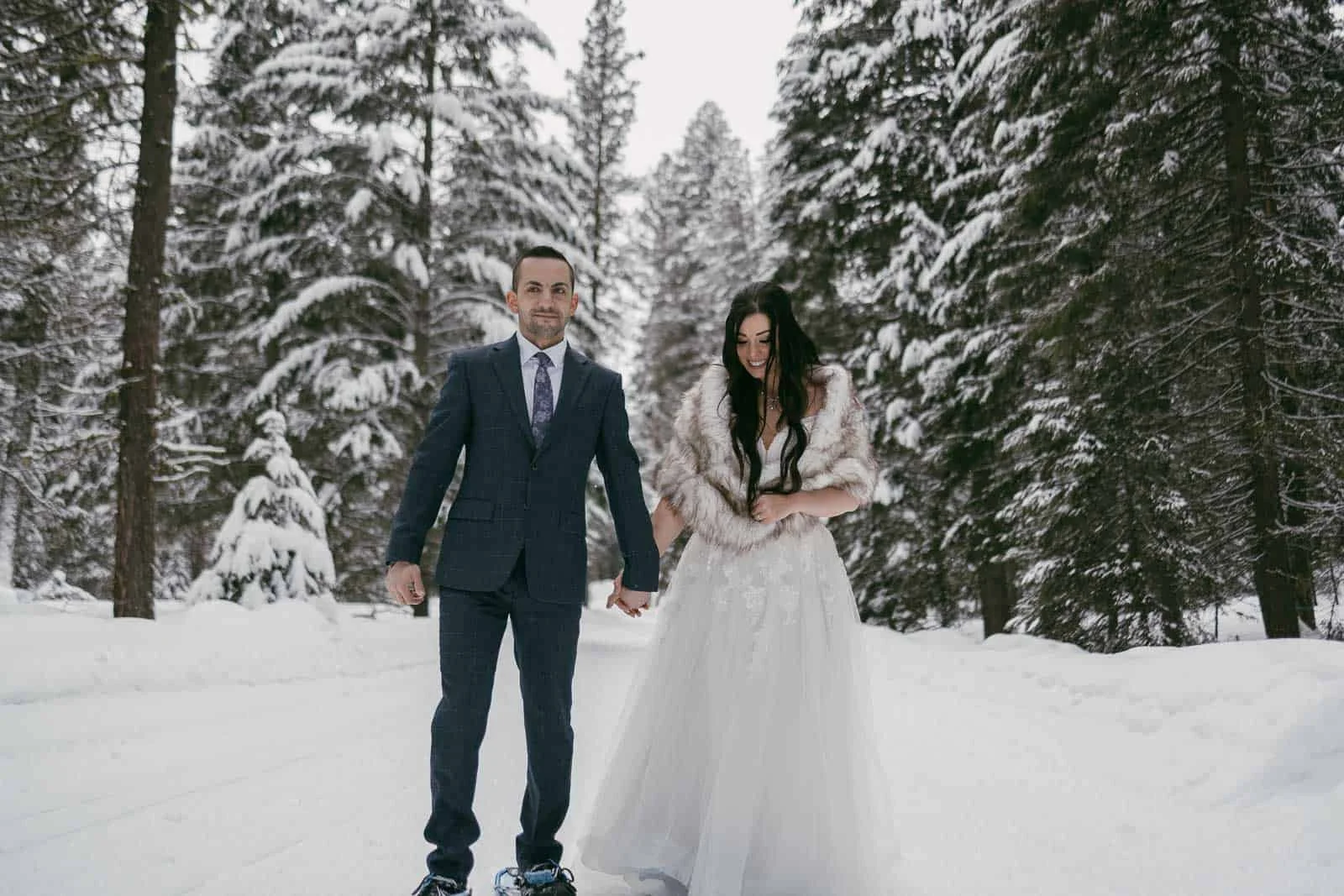  A couple dressed in elegant formal clothing stands hand in hand in a snow-filled forest setting. The man is wearing a suit paired with snowshoes, while the woman is in a long flowing dress layered with a fur shawl. They are surrounded by tall trees 