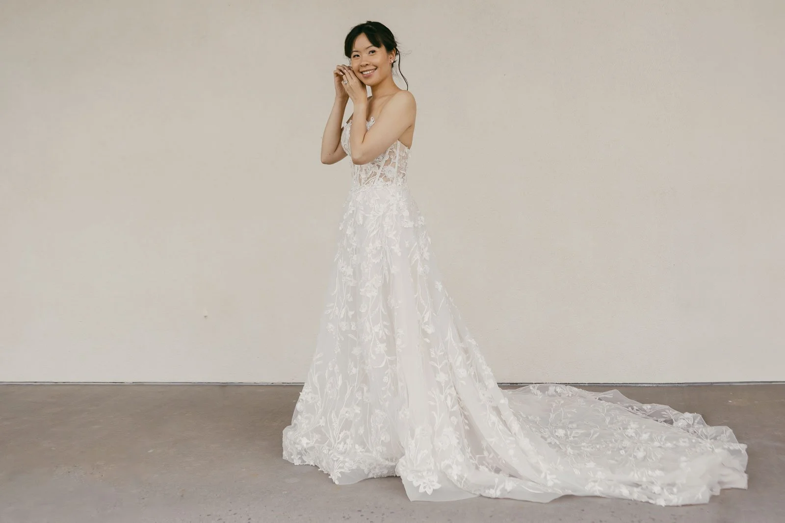 ride smiling over her shoulder in a strapless floral lace A-line gown with a dramatic cathedral train in a clean, minimalist studio setting