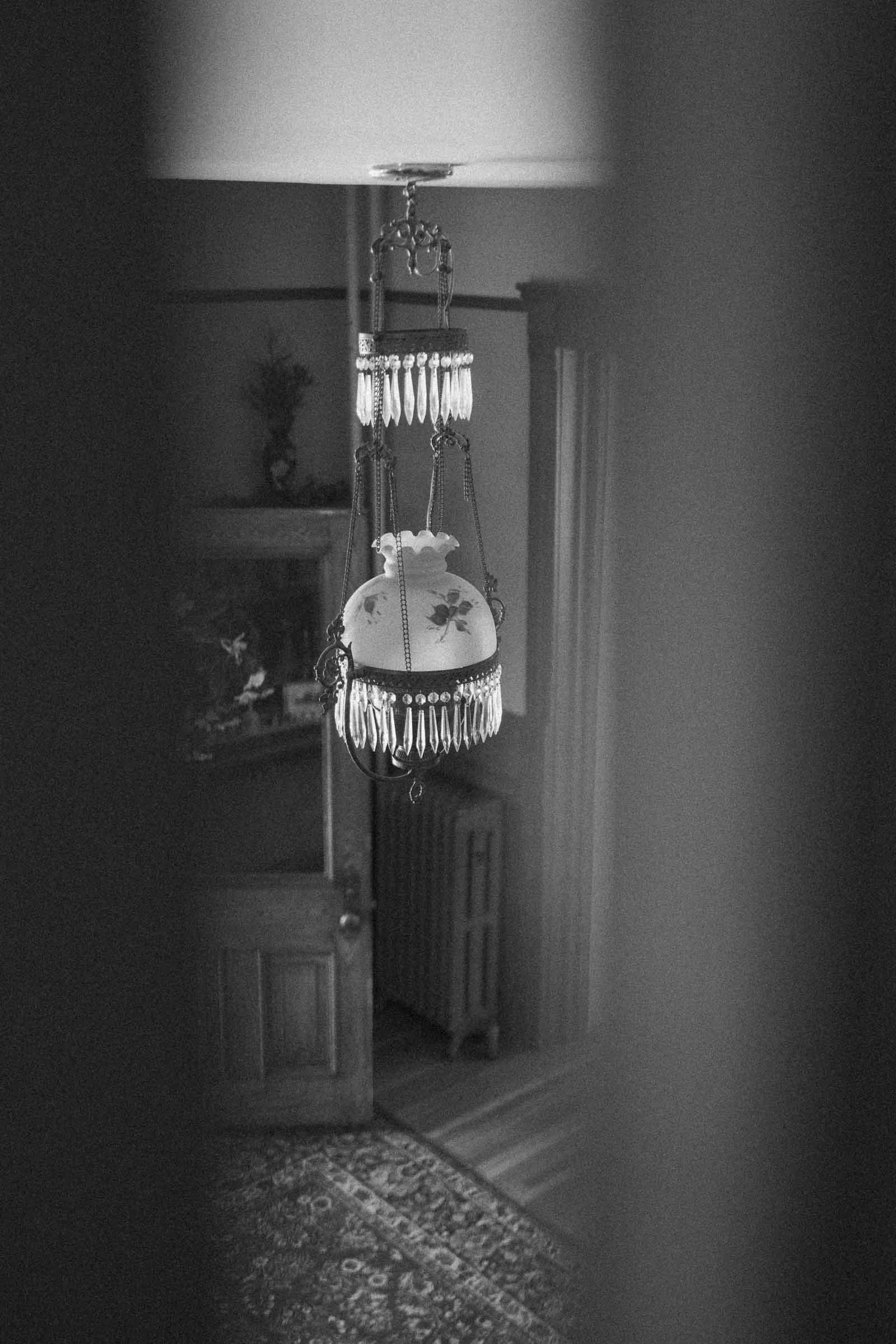  A vintage glass chandelier with floral patterns hangs from the ceiling in a dimly lit room with an open wooden door and patterned rug, seen through a narrow gap. Black and white photo. 