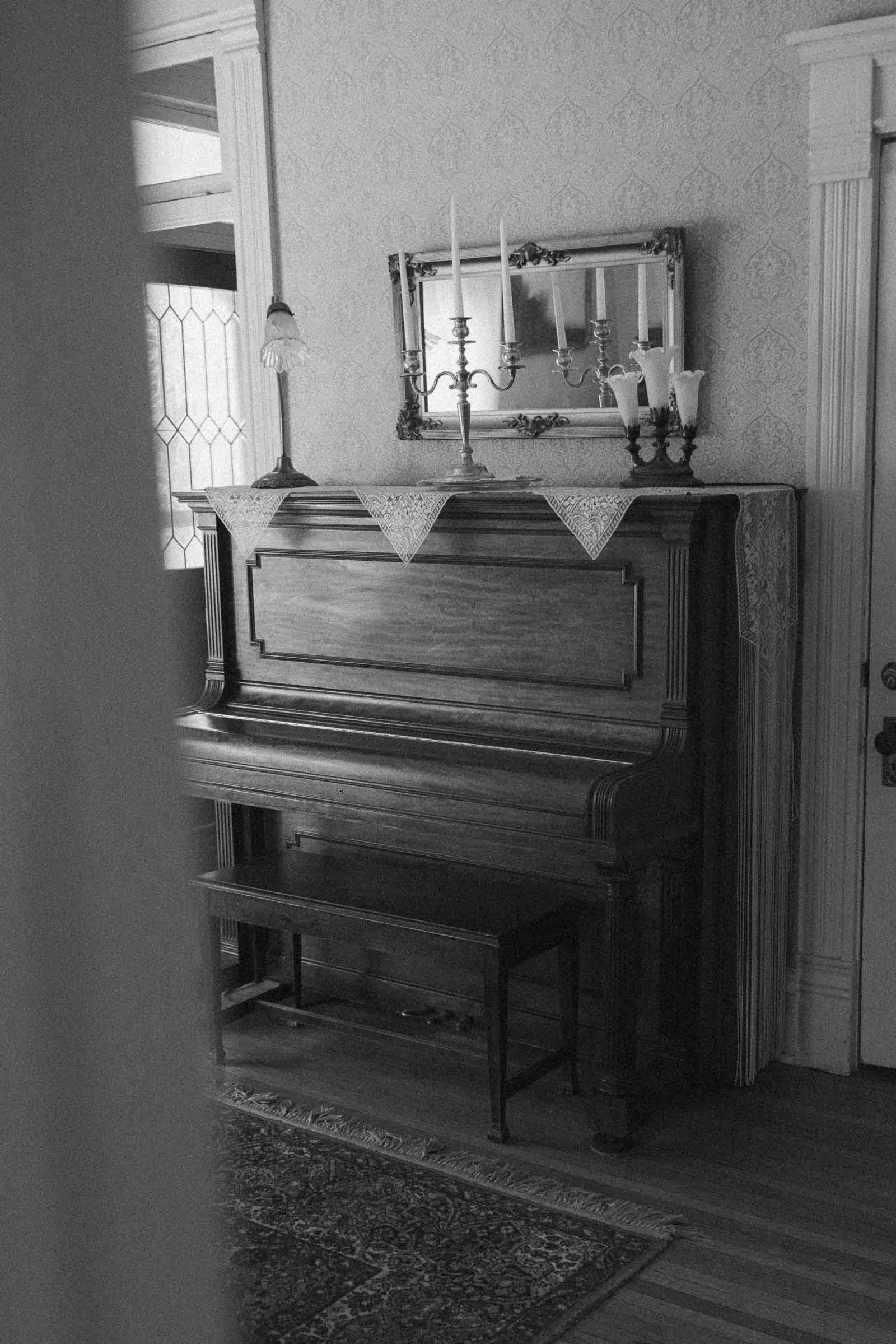  A black and white photo of an upright piano with a bench in front, a lace runner, a candelabra, and lamps on top. A mirror hangs above the piano, and patterned wallpaper decorates the wall behind. 