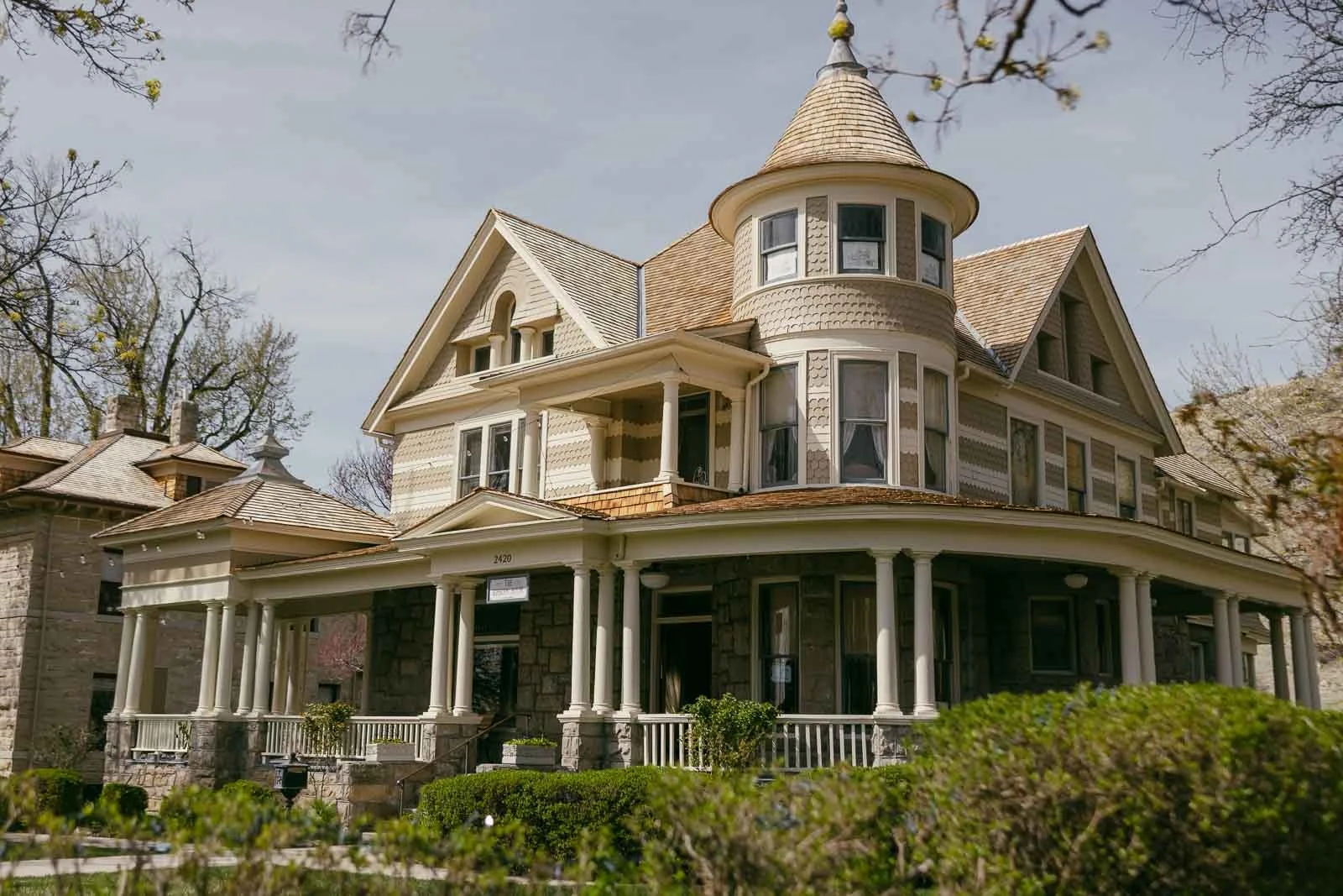  A large, historic Victorian house with a wraparound porch, detailed woodwork, a rounded turret, and beige siding, surrounded by greenery and trees under a partly cloudy sky. 