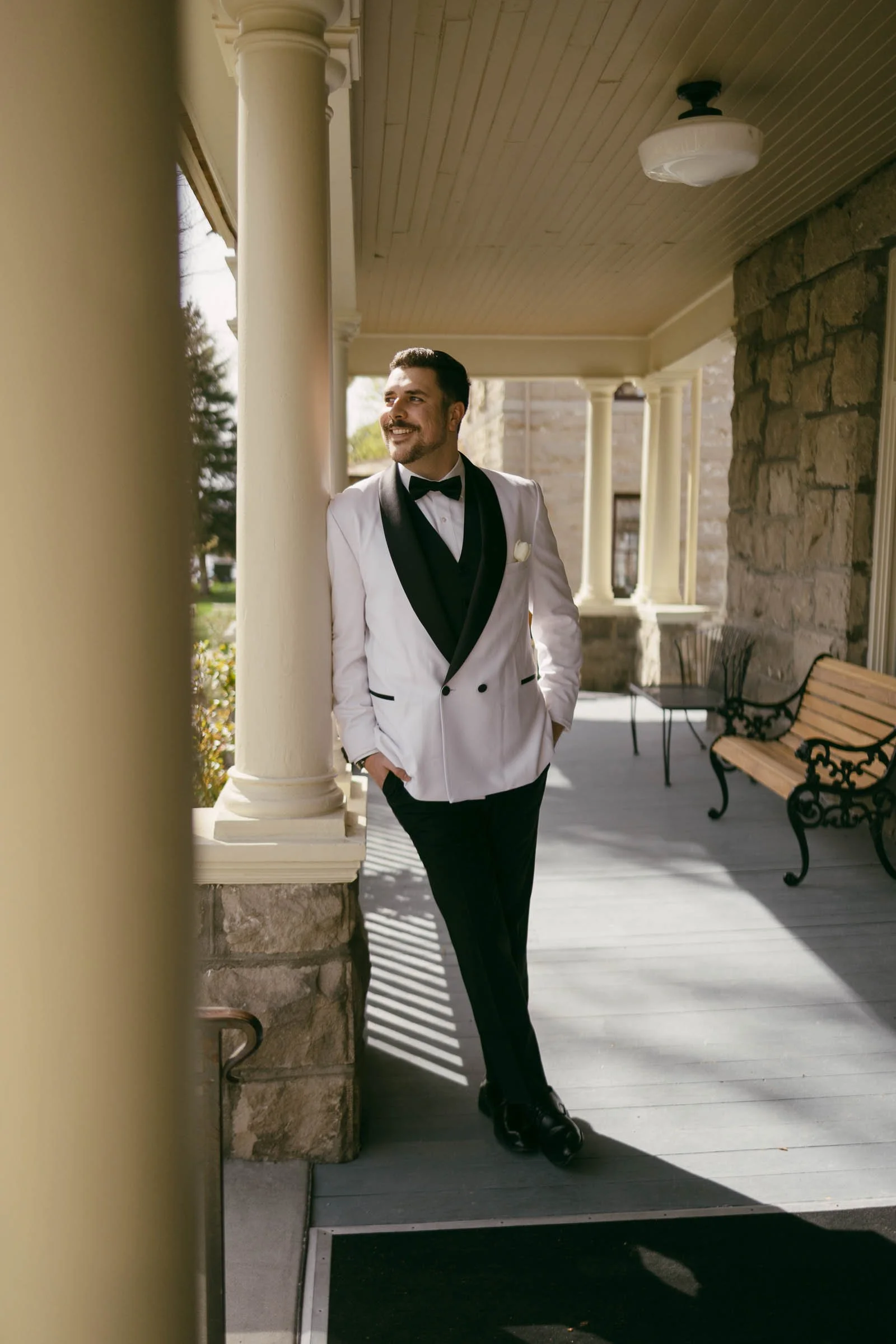  A man in a white tuxedo jacket and black pants stands smiling and leaning against a column on a porch with stone walls, a bench, and a light fixture above. 