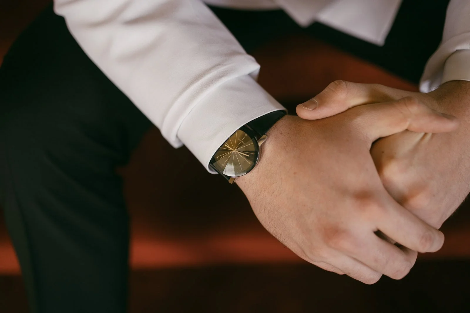  Close-up of a person wearing a white shirt and black pants, sitting with hands clasped. A black wristwatch with a gold face is visible on the left wrist. The background is blurred. 