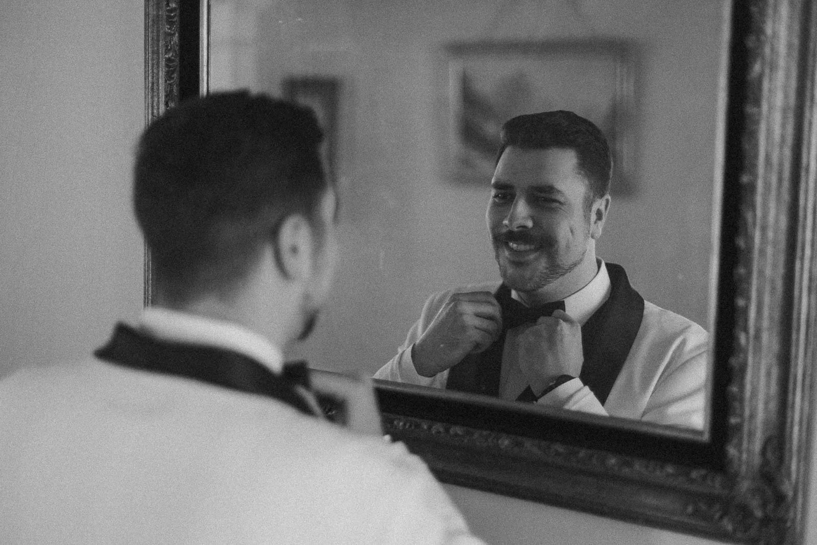  A man in a tuxedo adjusts his bow tie while smiling at his reflection in a framed mirror. The scene is in black and white, giving it a classic, timeless feel. 